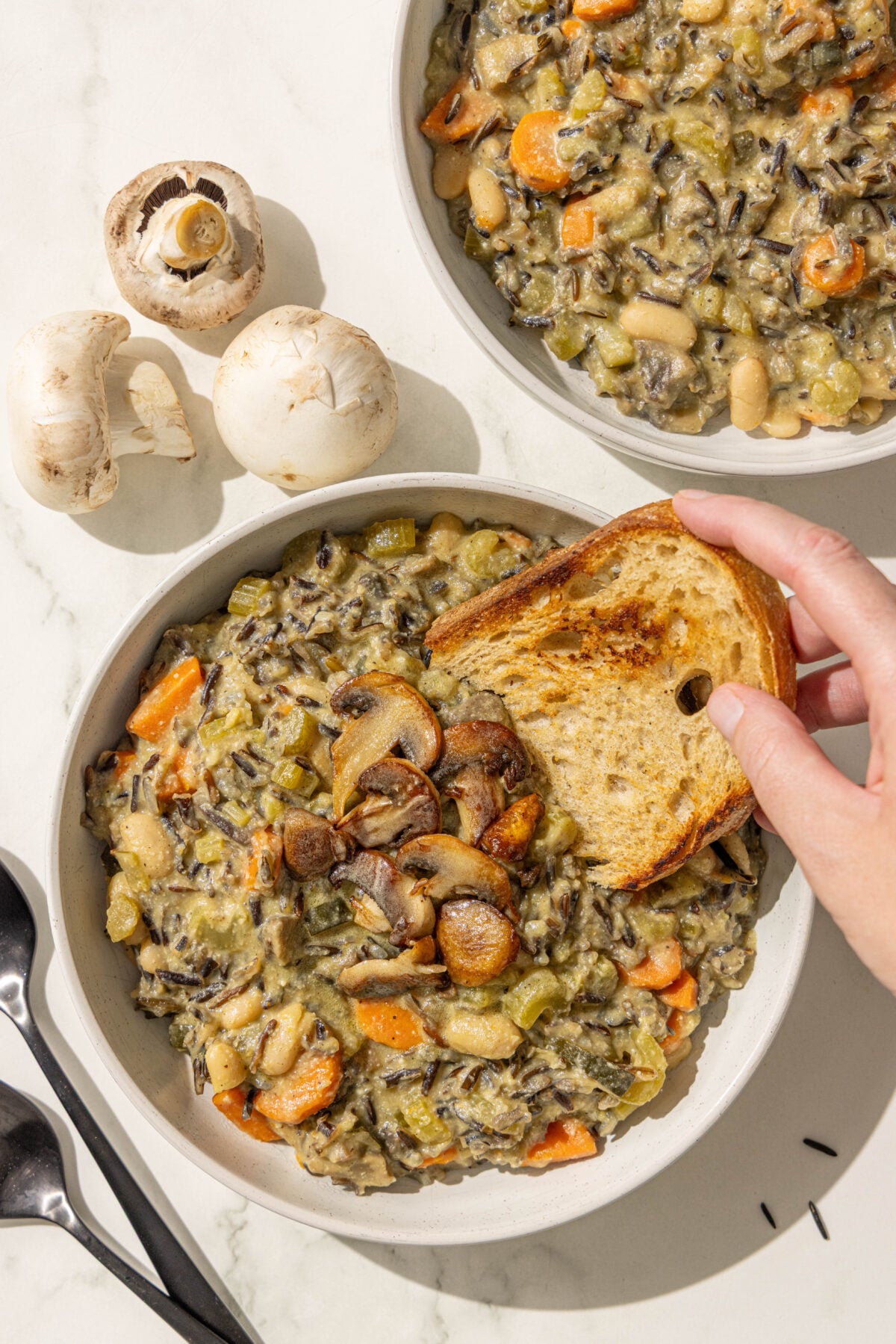Two bowls of creamy vegan wild rice soup topped with sauteed mushrooms and a white hand holding a piece of toast dipping into the soup.