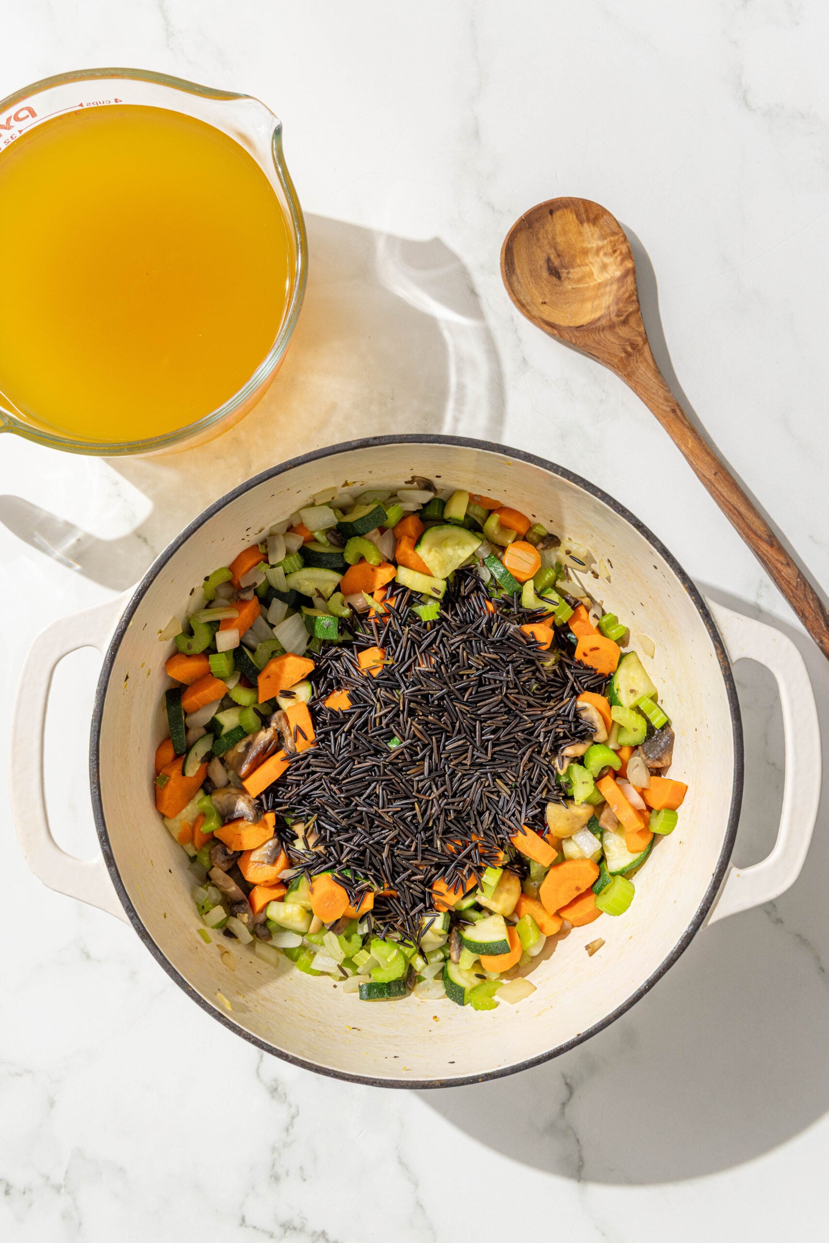 A large soup pot filled with sauteed carrots, onion, celery, and mushrooms topped with wild rice and then a large glass dish of vegetable broth and a wooden spoon.