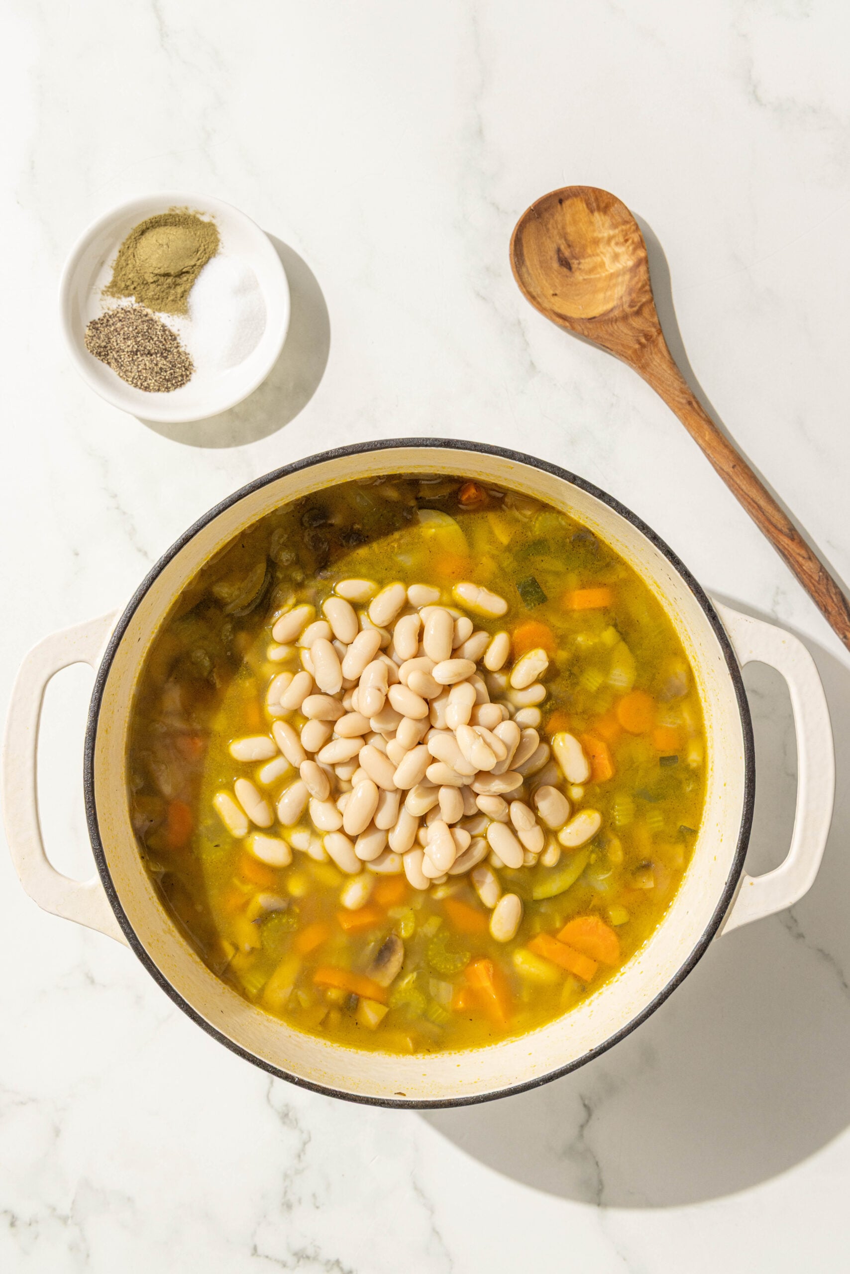 A large pot filled with soup topped with beans, a small bowl of spices, and a wooden spoon.
