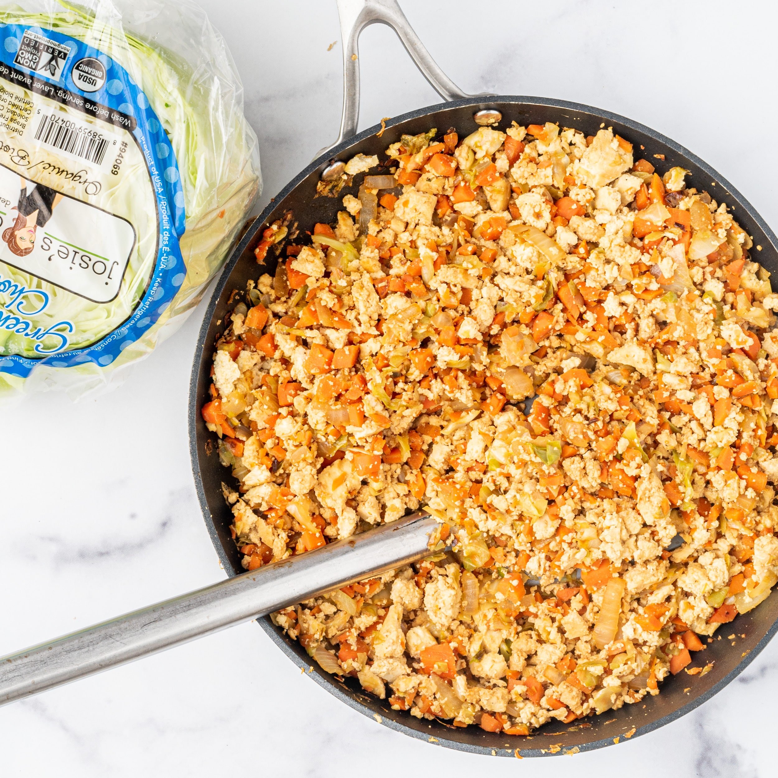 A large frying pan filled with cabbage, tofu, and carrots sautéed. Beside the pan is a head of cauliflower wrapped in plastic.