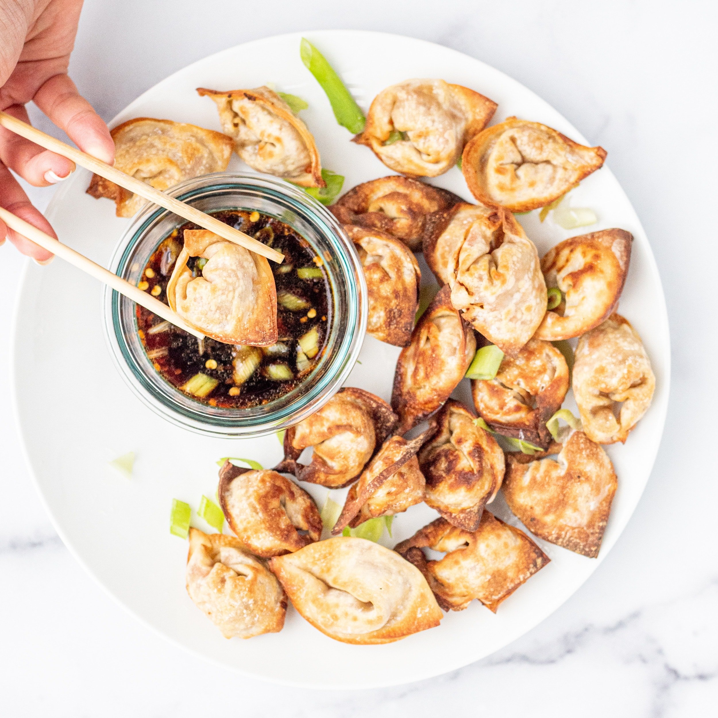 A birds eye view of a white plate filled with crispy wontons and a clear glass bowl of brown sauce with green onions on top. A white hand holds two chopsticks on the left and is dipping a wonton into the sauce.