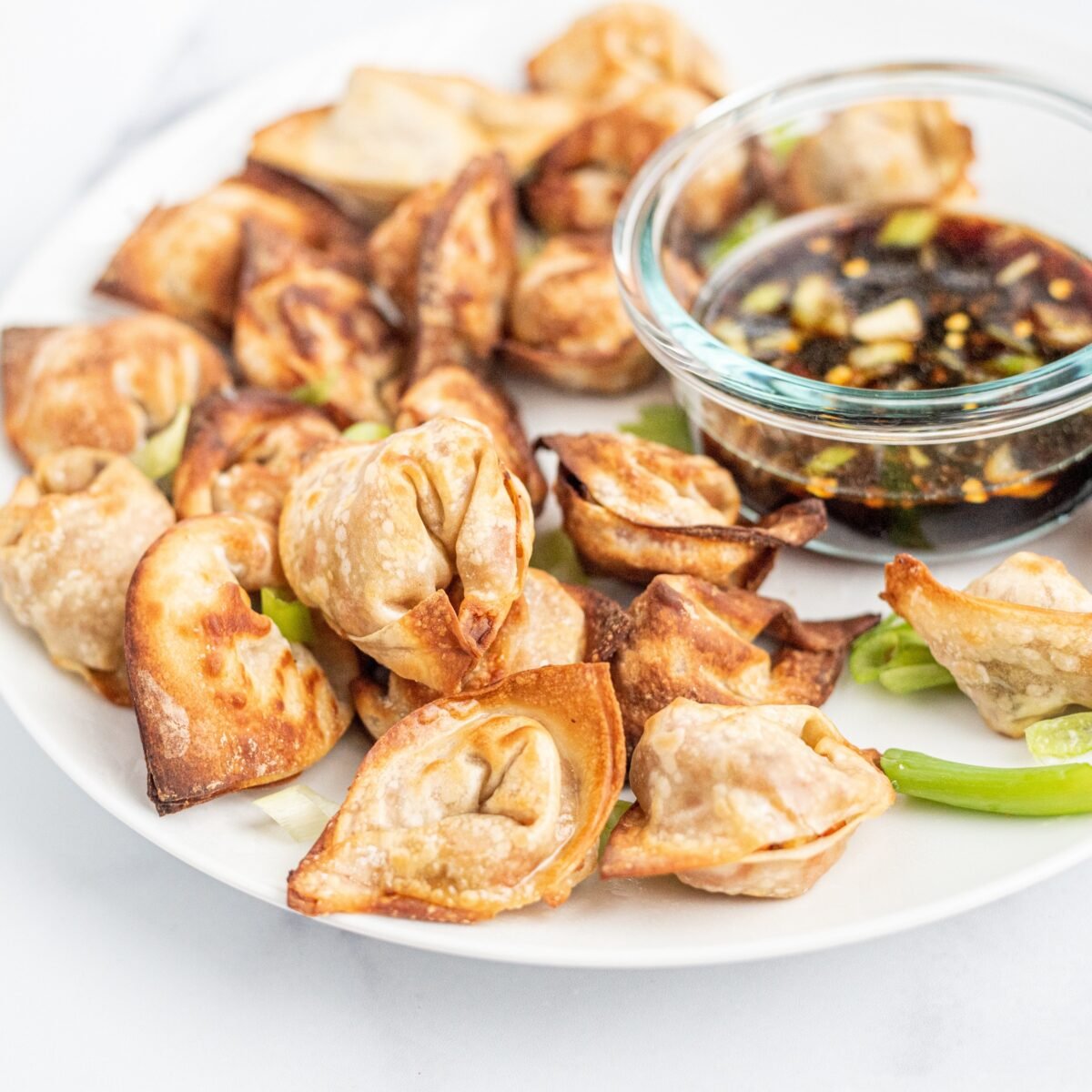 Crispy wontons on a white plate with green onions on top as garnish. On the right side of the plate is a clear glass bowl filled with a brown sauce and more green onions in it.
