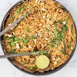A large pot filled with noodles garnished with cashews, fresh cilantro, and half a lime. In the center is a pair of tongs.