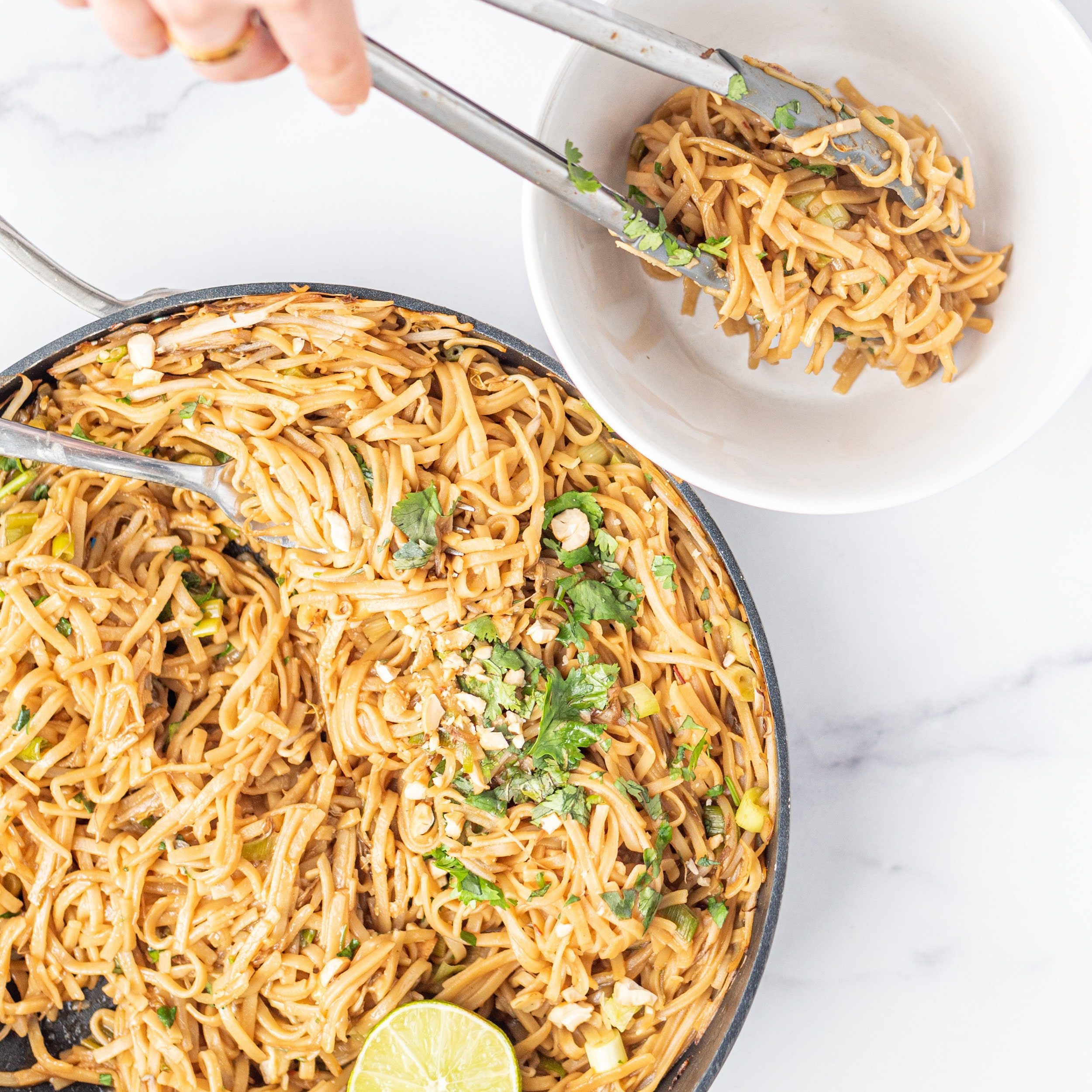 A large pan filled with thin noodles garnished with cashews, cilantro and lime. Beside the pan is a small white bowl with tongs in it placing a serving of noodles in it.