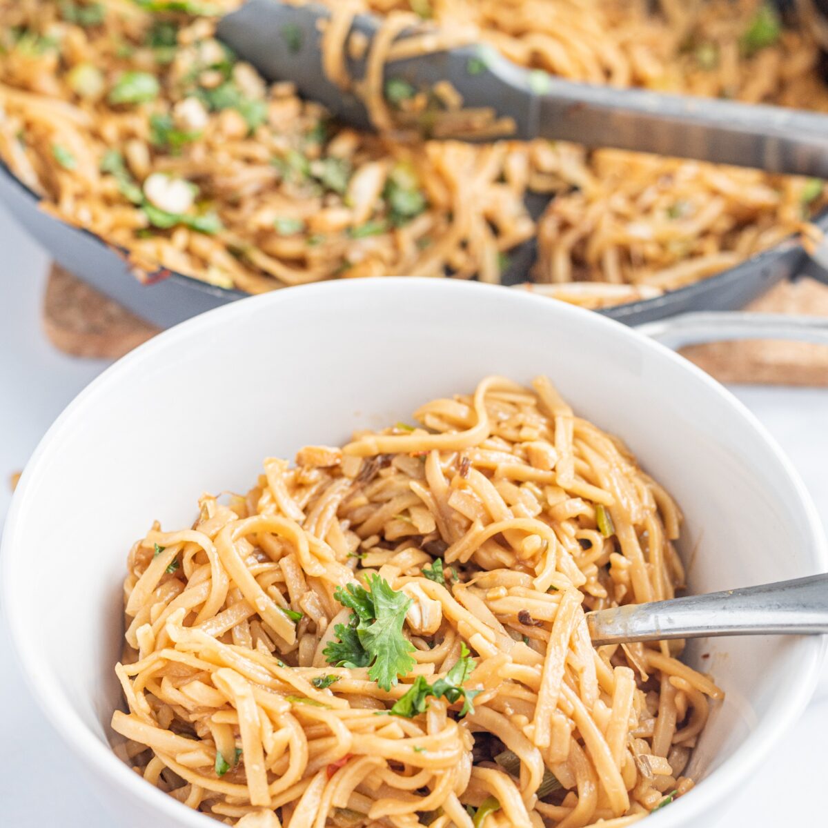 A white bowl filled with noodles with cilantro on top and a fork sticking out. Behind the bowl is a large pan with remaining noodles garnished with cilantro and lime with tongs in the pot.