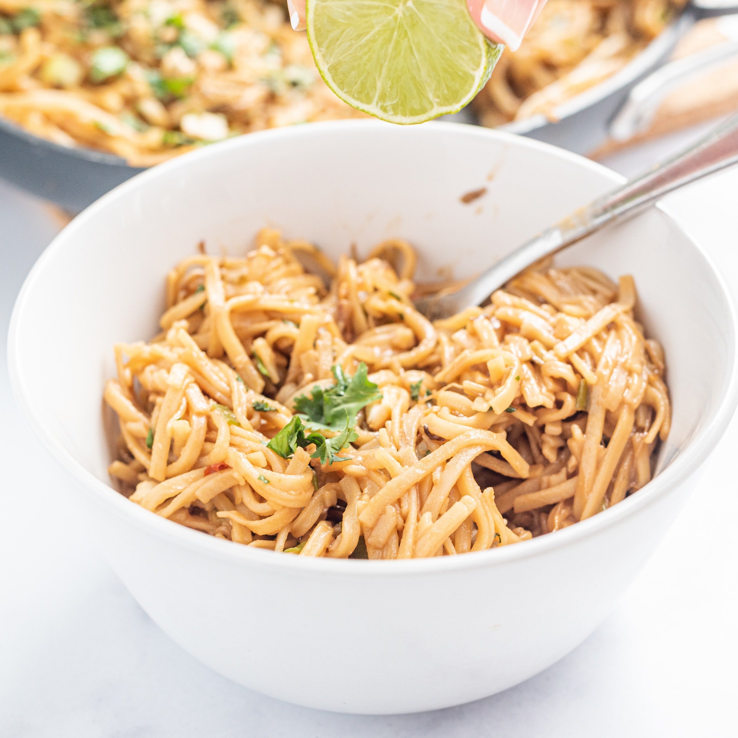 A white bowl filled with thin noodles with a silver fork sticking out and a lime over top being squeezed by a white hand. In the background is a large pan with remaining noodles.