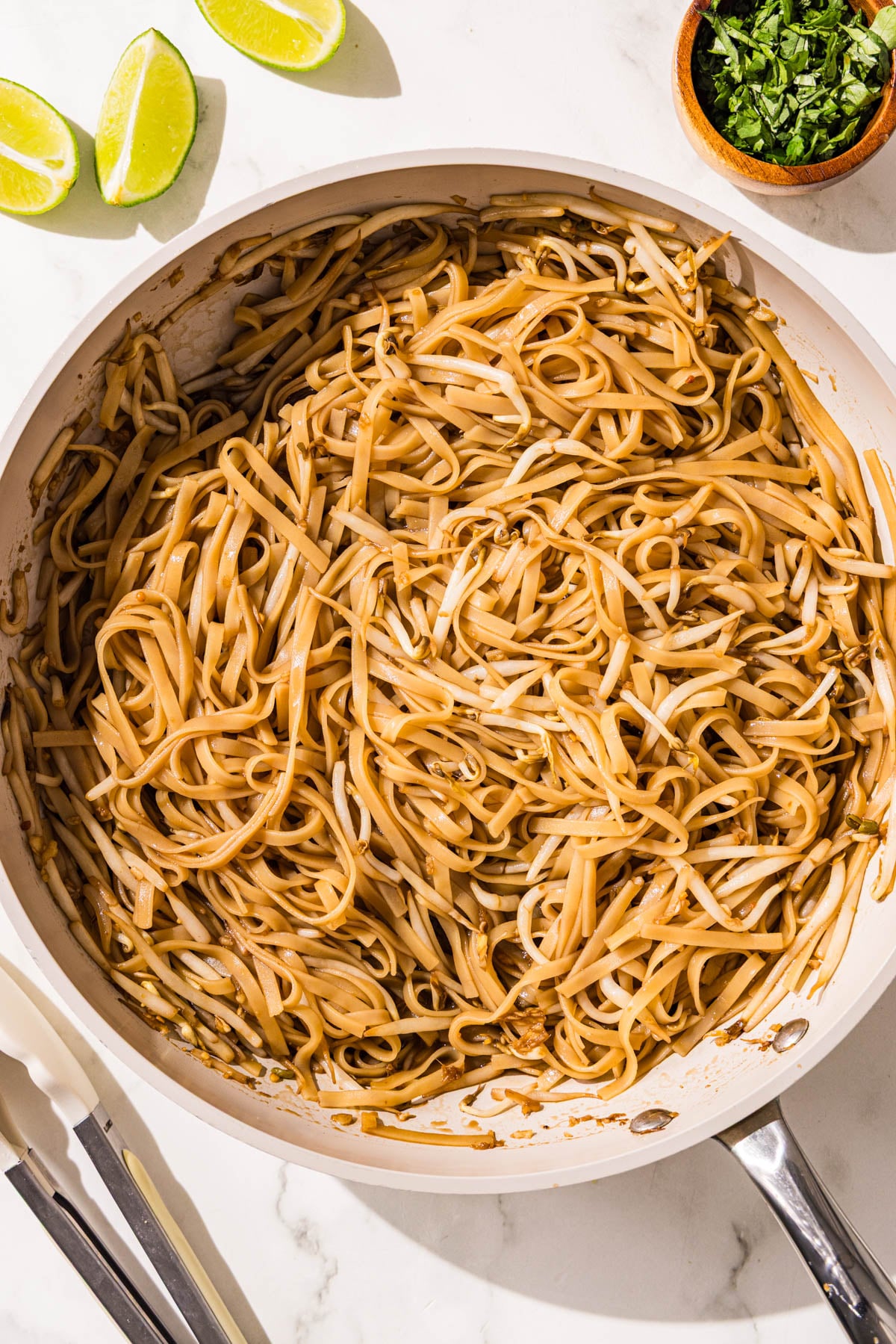A frying pan filled with saucy rice noodles and bean sprouts. Besides the pan is a pair of tongs, a small bowl of green onion, and 3 lime wedges.