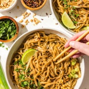 Two bowls of pad thai with chopsticks and each garnished with a lime wedge, fresh cilantro, peanuts and green onion, and a white hand in one bowl holding the chopsticks grabbing a bite. Beside is three more bowls of chopped peanuts, cilantro, and soy sauce plus three lime wedges on the counter.