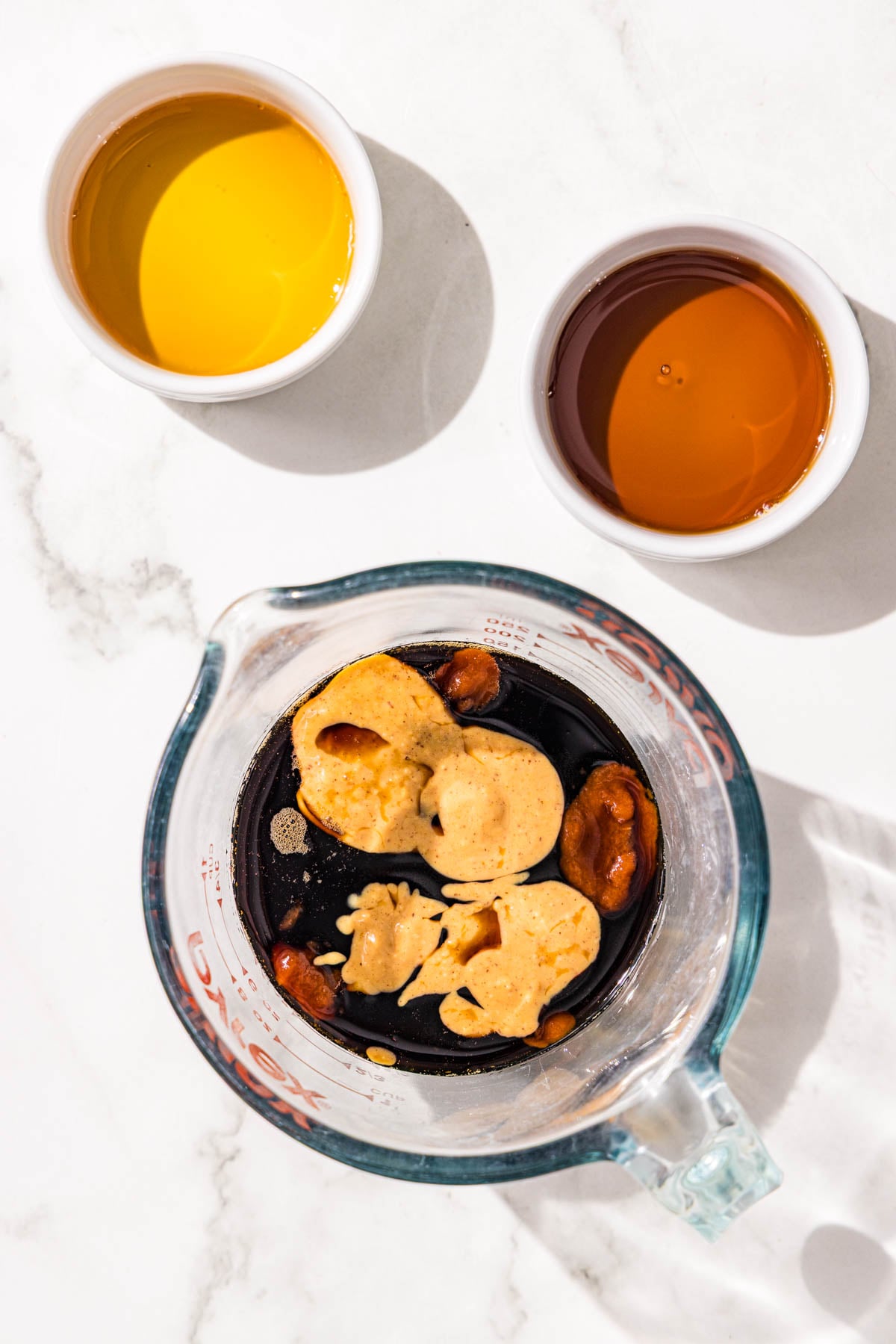 A glass jar filled with coconut aminos and peanut butter plus two small white glass bowls of maple syrup and sesame oil.