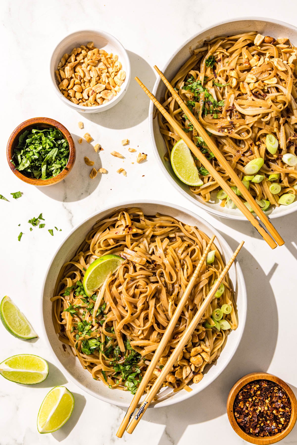 Two bowls of pad thai with chopsticks and each garnished with a lime wedge, fresh cilantro, oeanuts and green onion. Beside is three more bowls of chopped peanuts, cilantro, and soy sauce plus three lime wedges on the counter.
