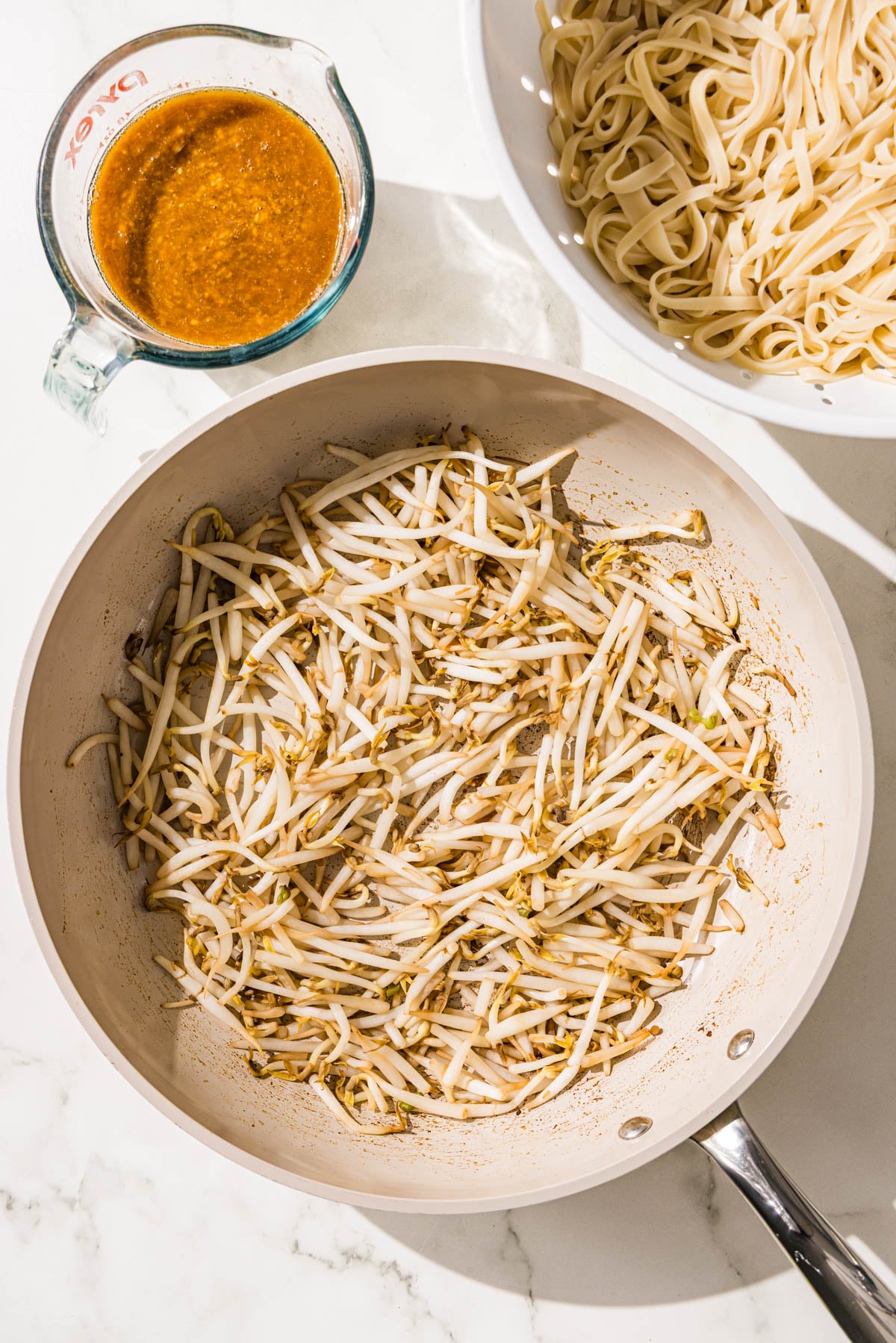 A pan filled with bean sprouts, a jar of sauce, and a colander filled with noodles.