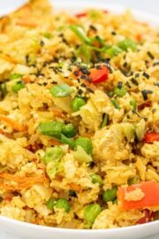 A white bowl filled with yellow rice mixed with veggies with sesame seeds on top and a pair of chopsticks sitting out the side of the bowl.