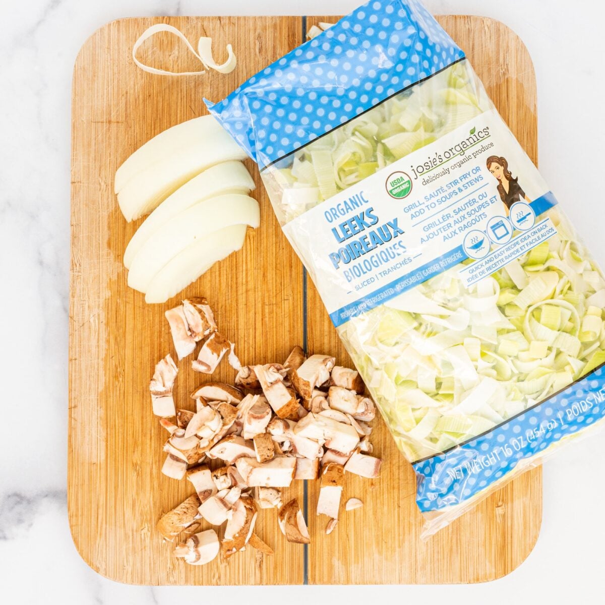 A wooden cutting board with an onion cut in slices, chopped mushrooms, and a bag of organic sliced leeks.