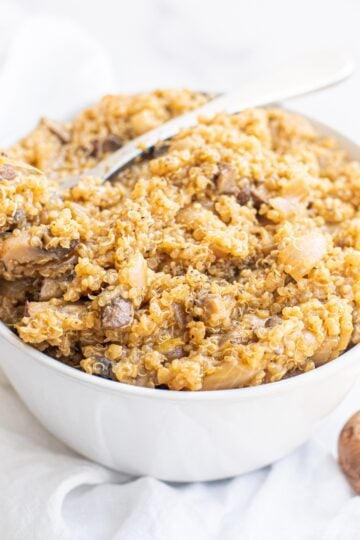 A bowl of Vegan One Pot Mushroom Quinoa Risotto on a white cloth towel with two mushrooms sitting on the counter beside the bowl.