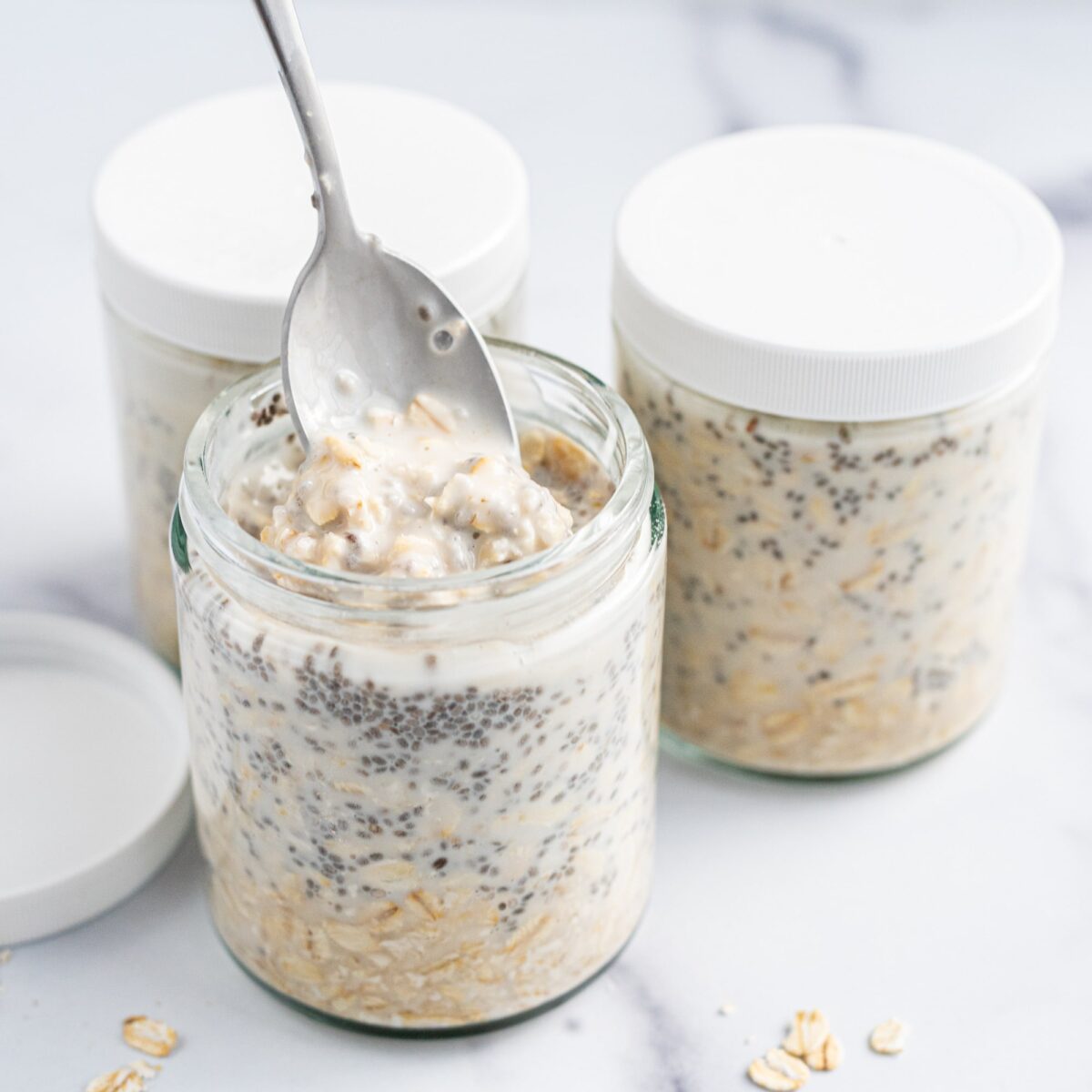 Three jars of Vegan Chia Overnight Oats. The back two jars have lids on them while the front jar's lid is off and to the side and a silver spoon is in the oats.