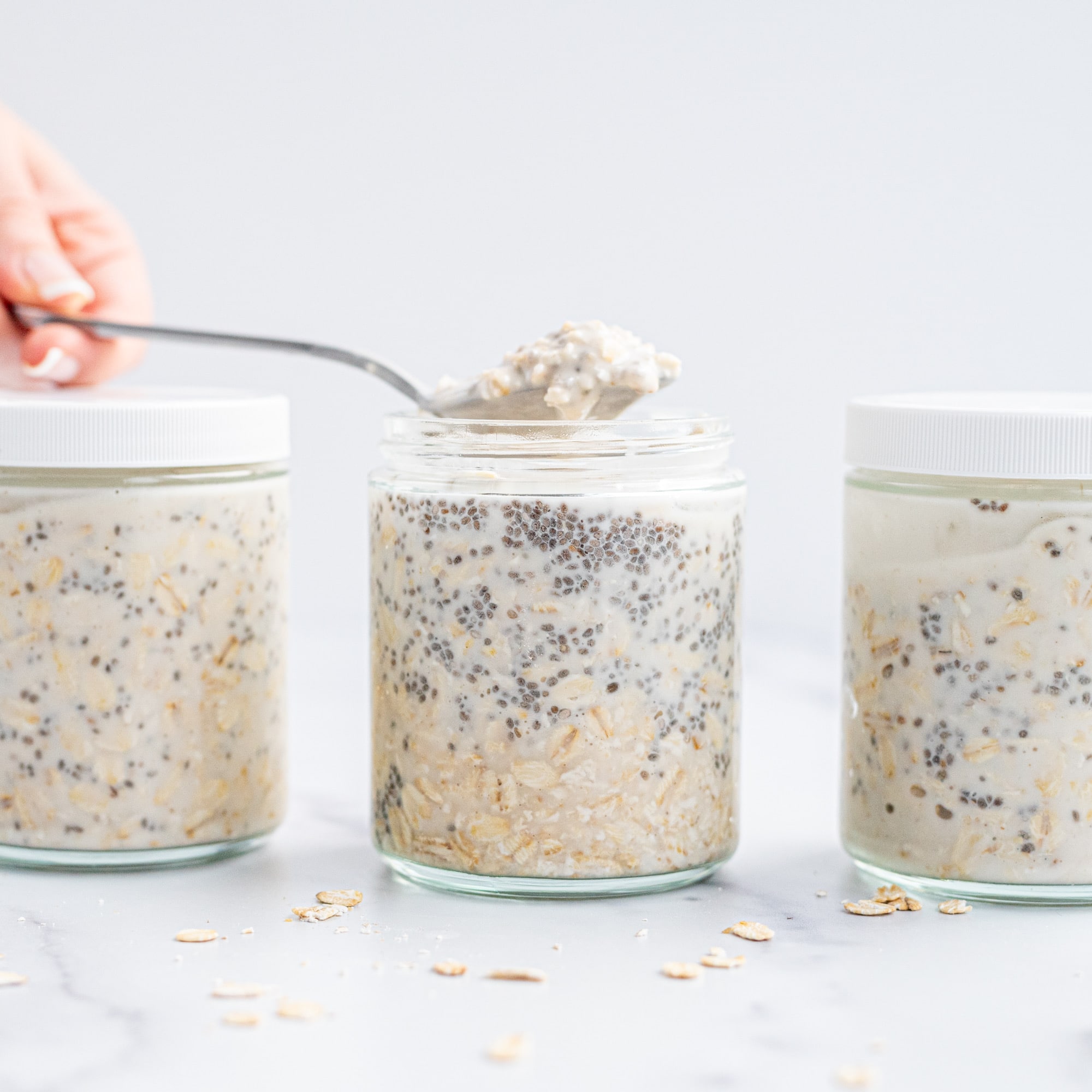 Three jars of vegan chia overnight oats. The two outer jars have lids on them and the center jar has a silver spoon sticking out of it with a scoop of oats on it with a white hand holding the spoon from the left of the image.
