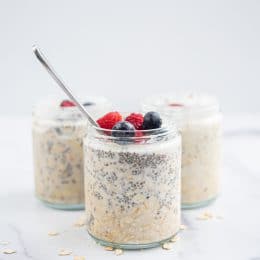 Three glass jars of Vegan Chia Overnight Oats topped with blueberries and raspberries. The jar in the front has a silver spoon sticking out of it. There are oats on the counter scattered around the jars.