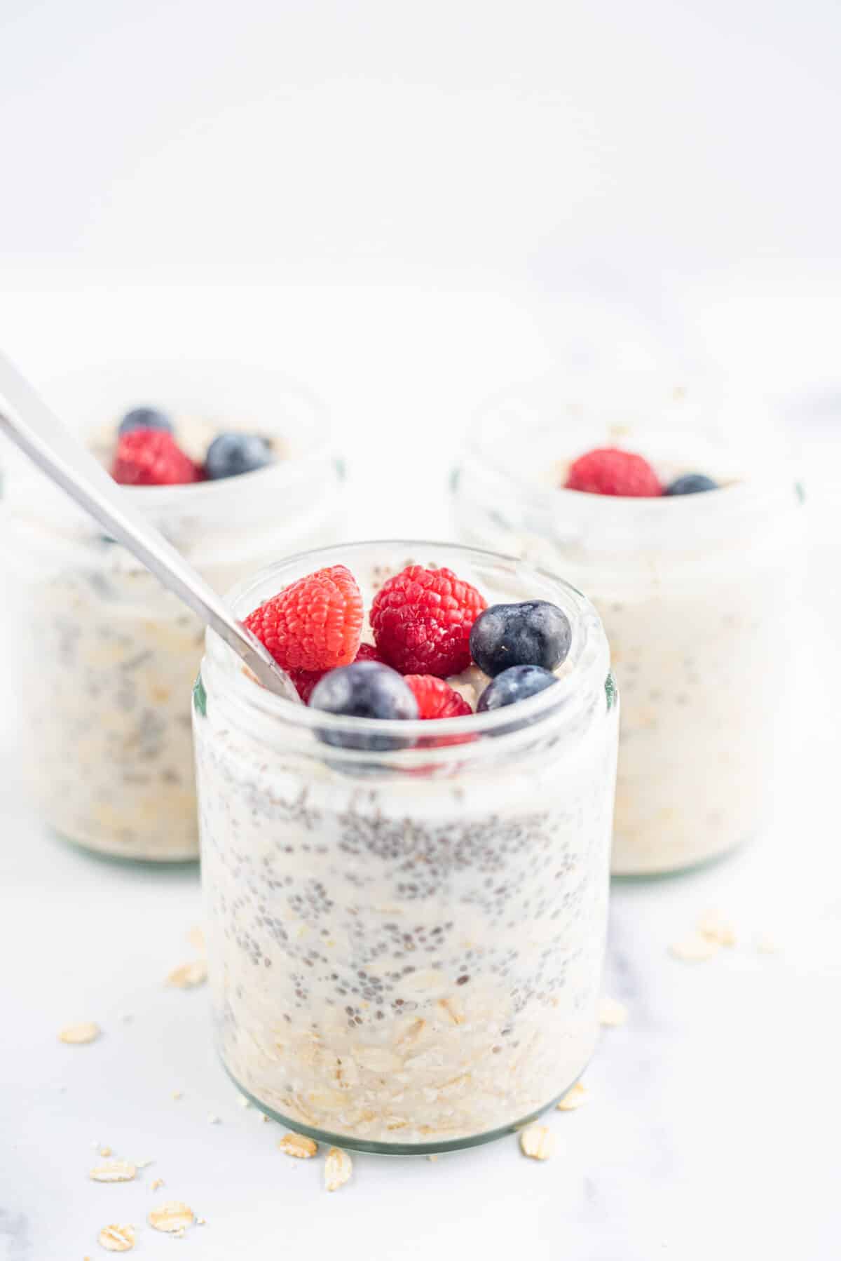 Three glass jars of Vegan Chia Overnight Oats topped with blueberries and raspberries. The jar in the front has a silver spoon sticking out of it. There are oats on the counter scattered around the jars.