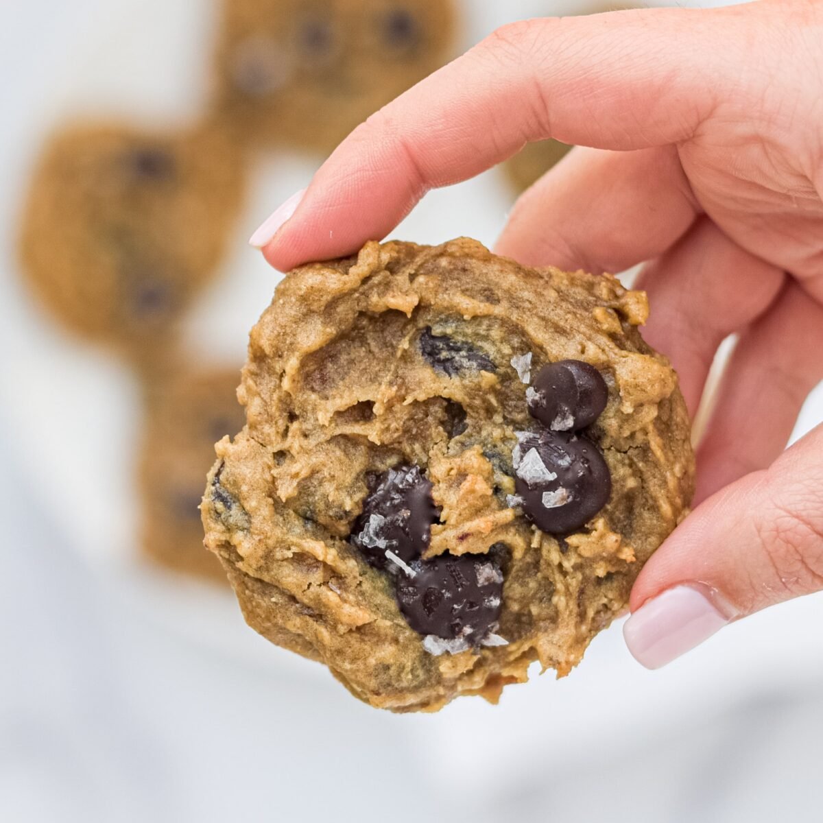 Close up of one Vegan Chickpea Chocolate Chip Cookie being held by a white hand with pink painted nails. In the background is a plate of cookies.