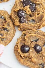 Vegan Chickpea Chocolate Chip Cookies on a white plate with a white hand with pink painted nails in the bottom left corner grabing a cooking off the plate.