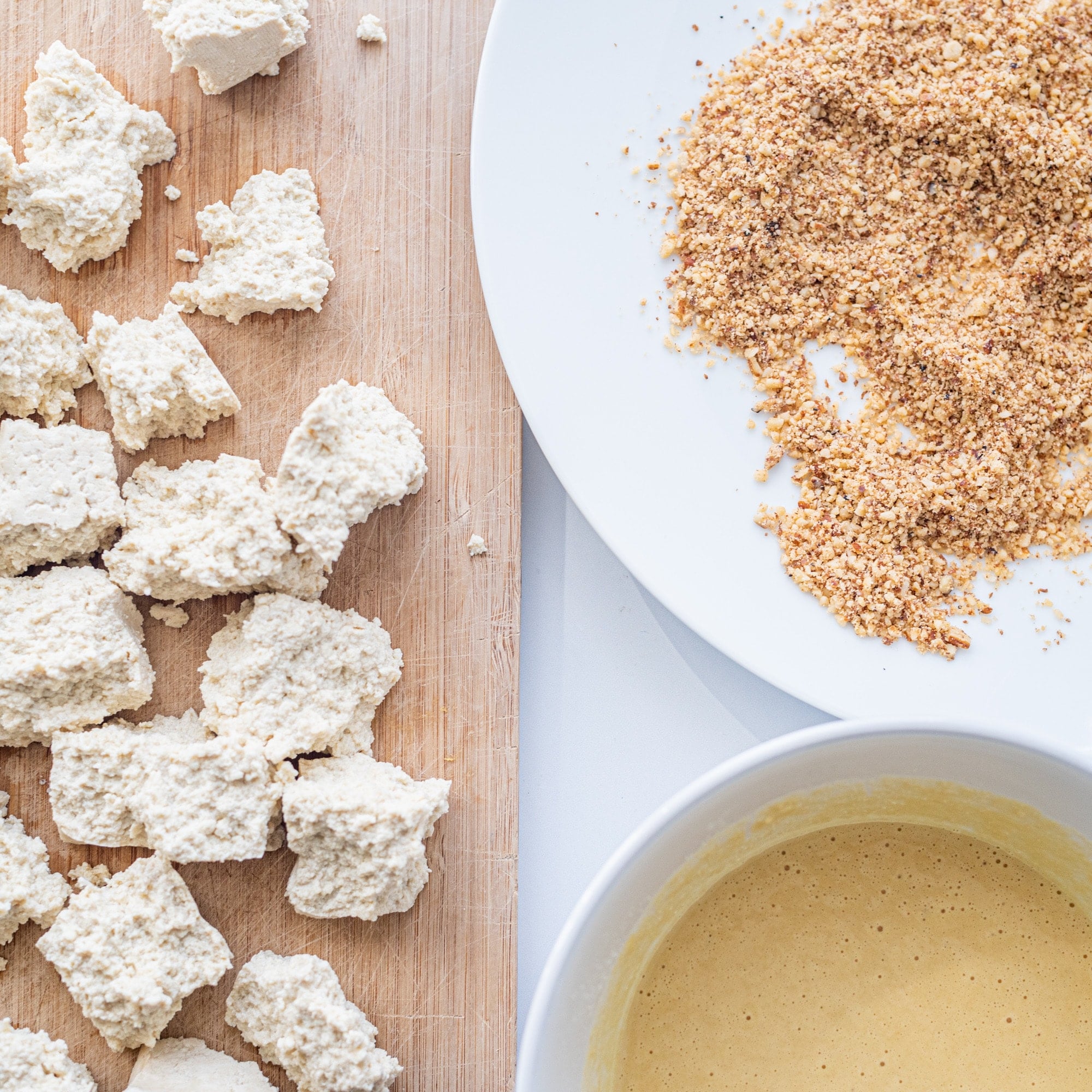 A cutting board with tofu pieces on it, a white plate with breadcrumbs, and a bowl of yellow batter.