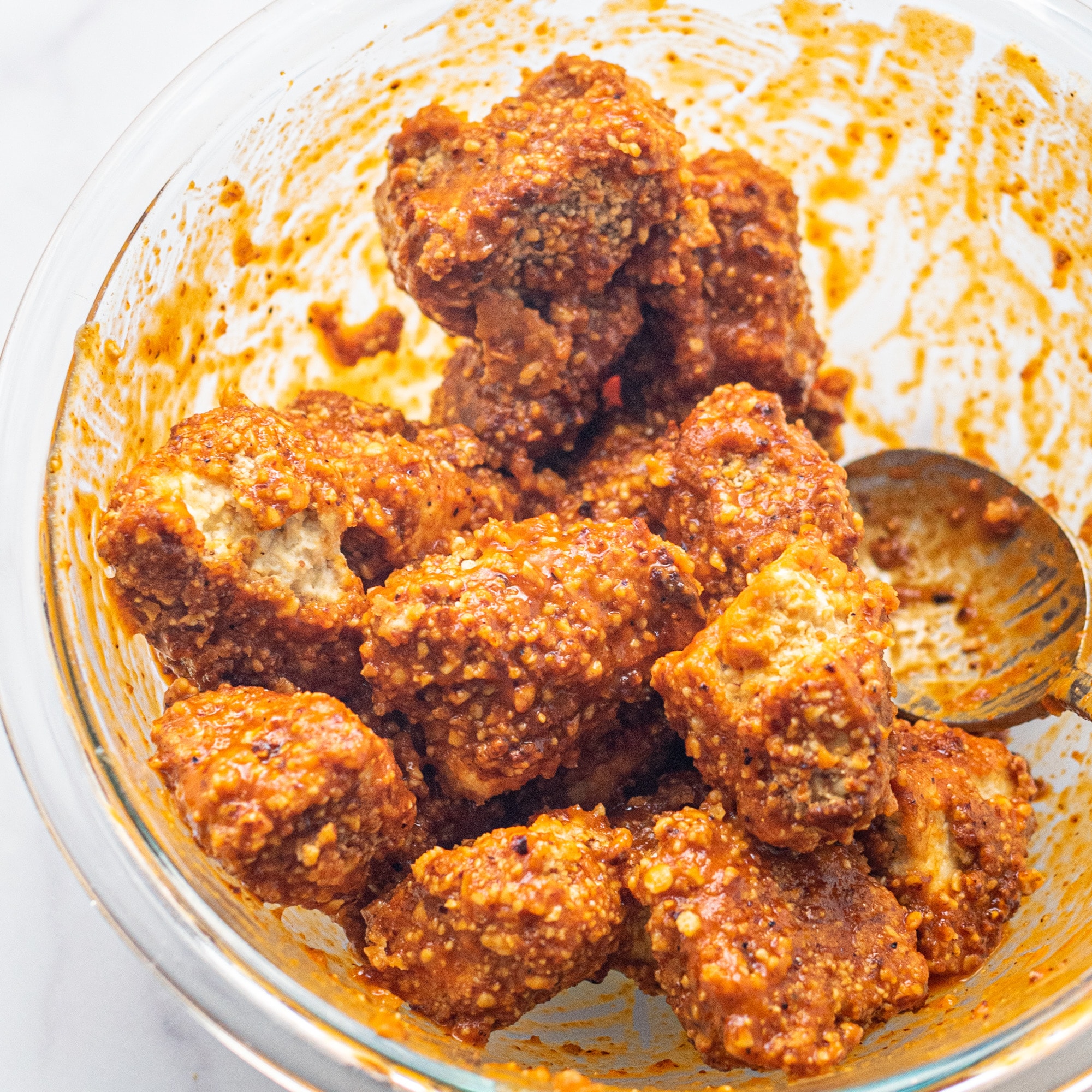 A glass bowl filled with tofu poppers in them tossed in buffalo sauce and a wooden spoon.