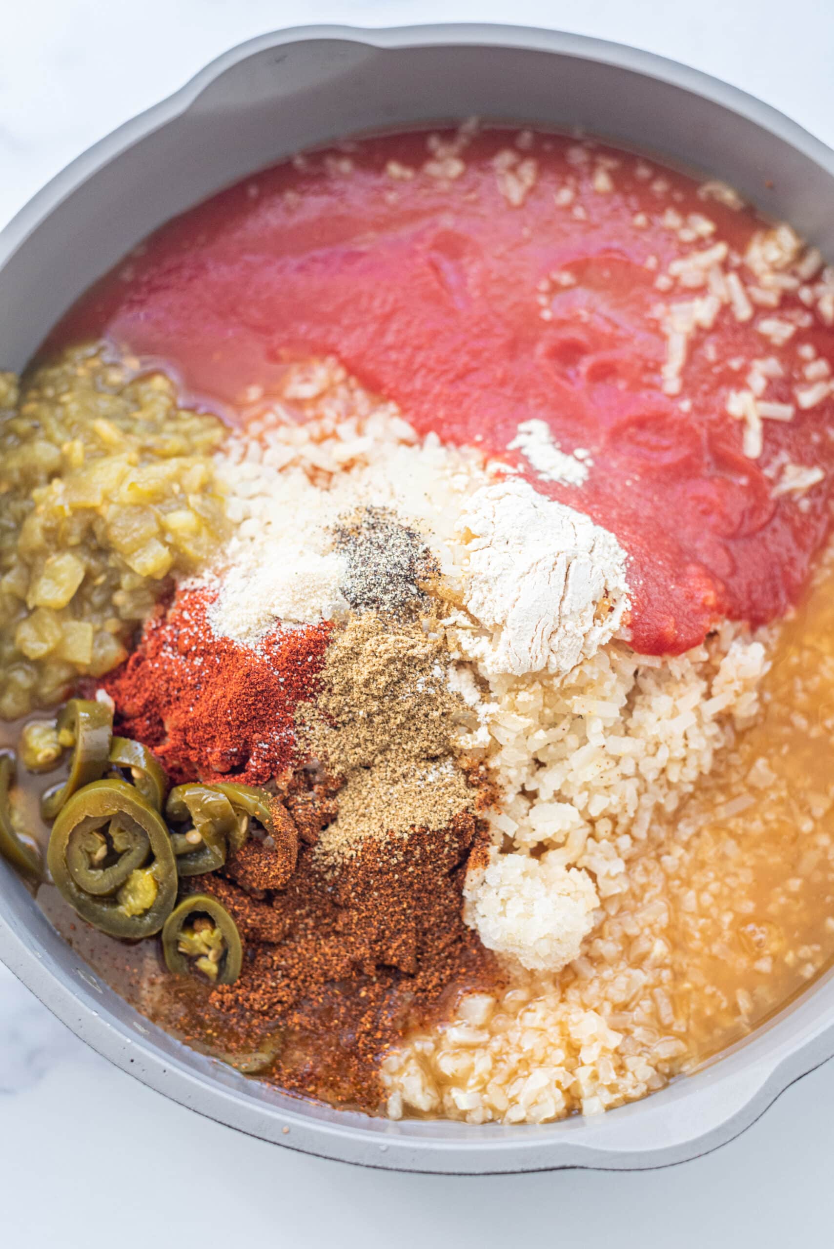 One Pan Creamy Mexican Cauliflower Rice ingredients (including tomato sauce, cauliflower rice, jalapenos, garlic, salt, paprika, cumin, mild green chiles, veggie broth) in a large pan ready to be stirred and cooked.