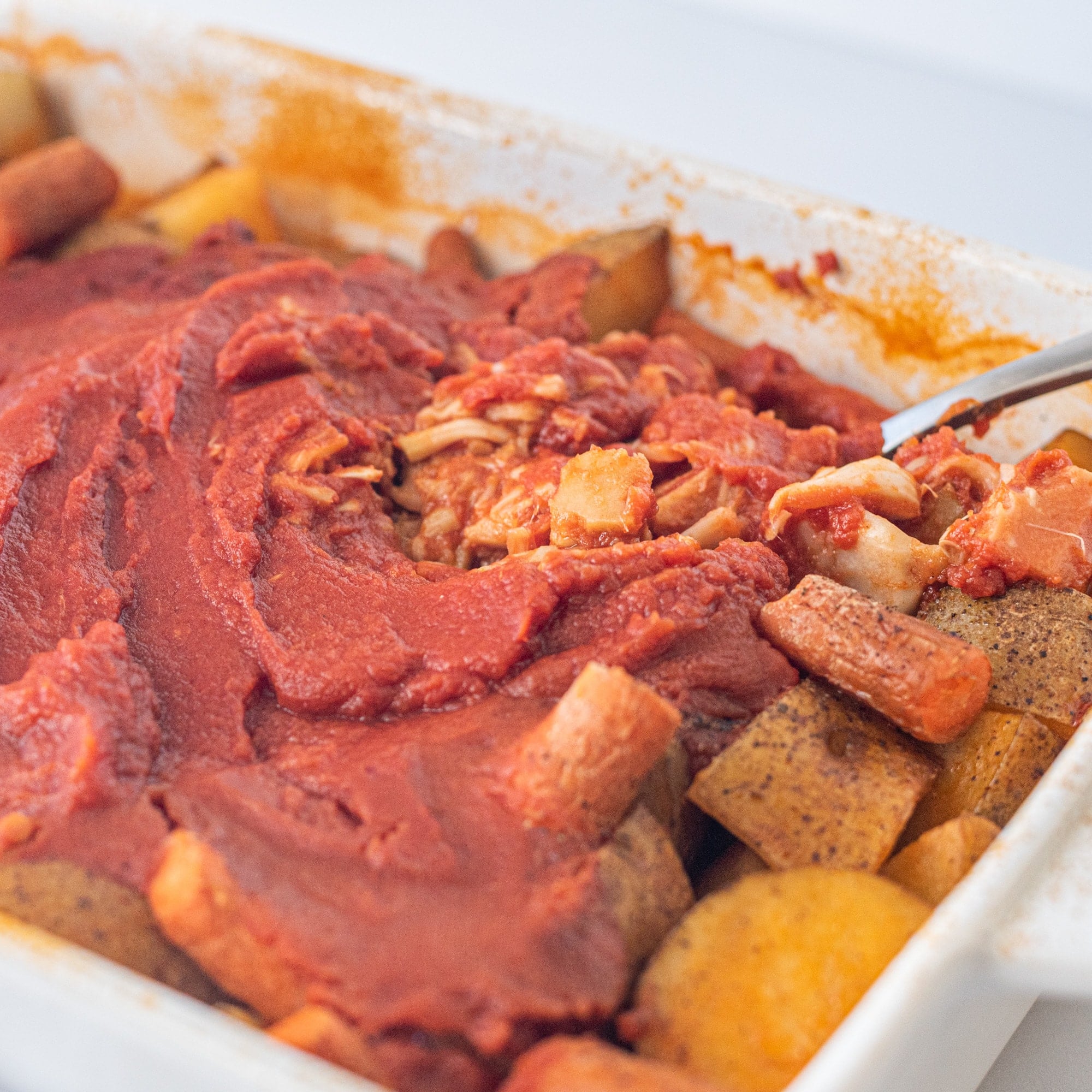 A casserole dish filled with potatoes, carrots, jackfruit, and a tomato sauce and a spoon.