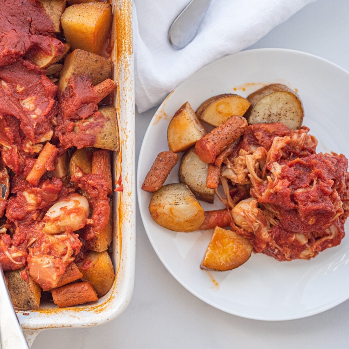 A plate with carrots, potatoes, and jackfruit brisket on in covered in a tomato sauce and a casserole dish with the rest of the dish.