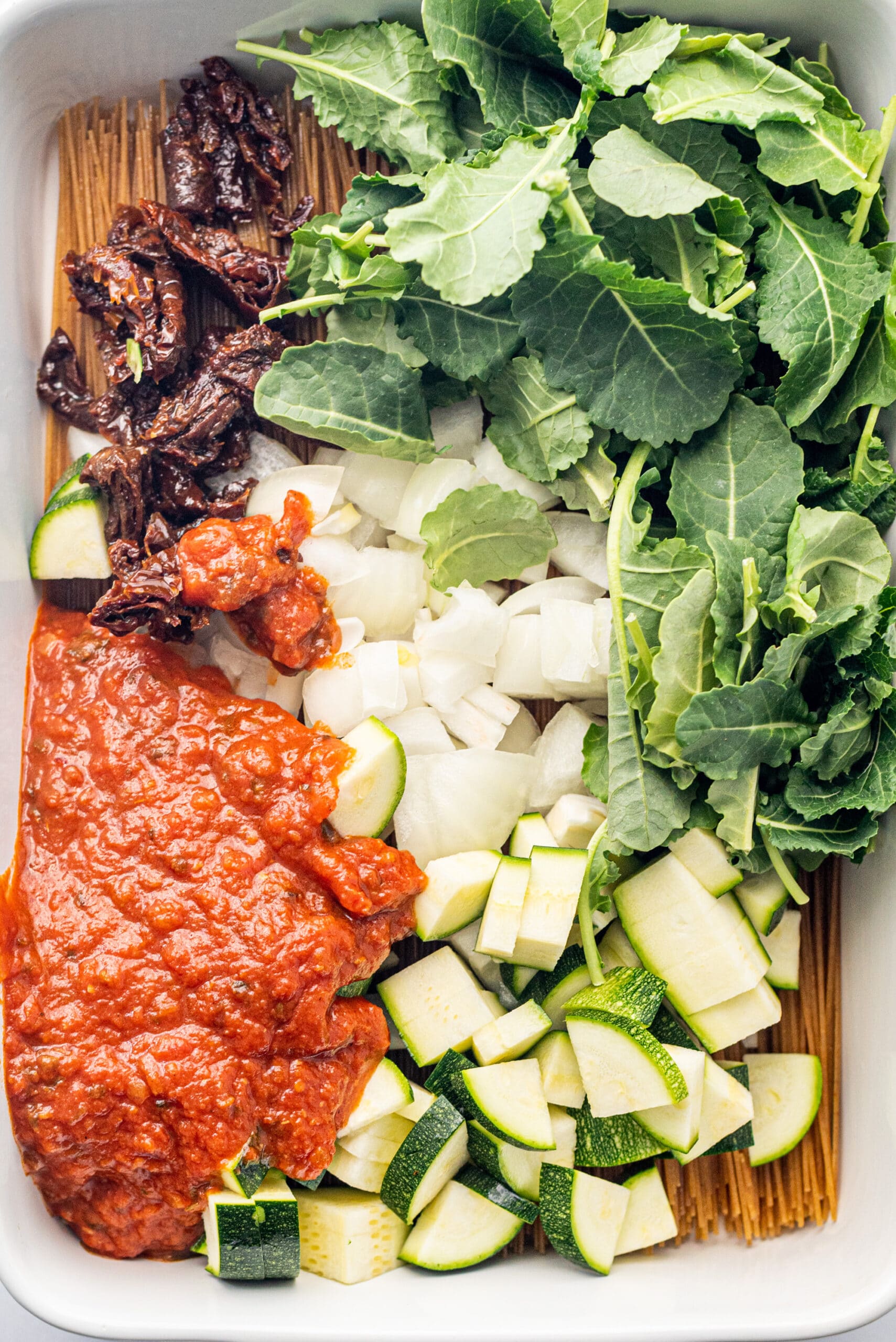 A 9x13 white glass casserole dish filled with dry spaghetti noodles topped with a pile of kale, chopped zuchinni, sundried tomatoes, diced onion, and marinara.