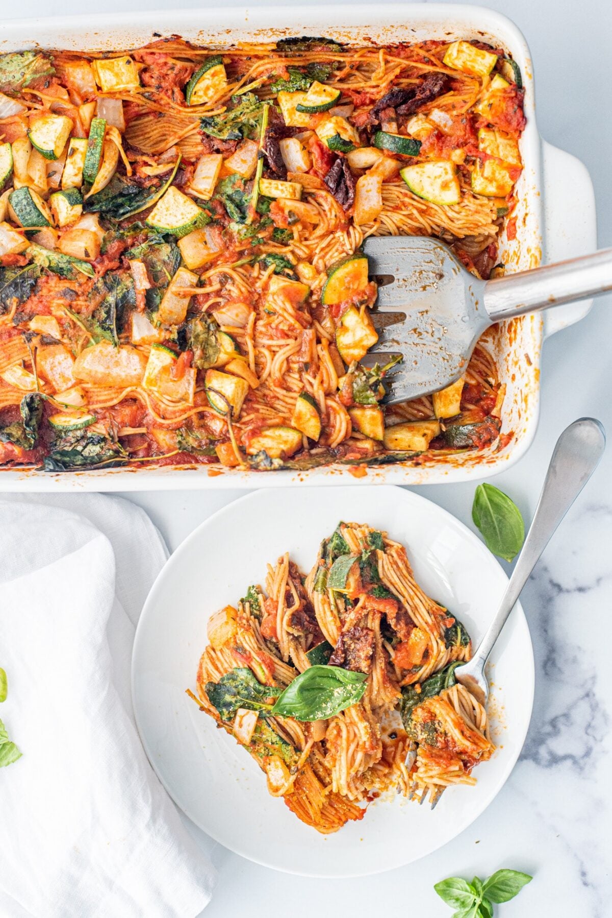 A casserole filled with baked spaghetti, a grey silicone spatula in the spaghetti, and a small white plate with a scoop of spaghetti on it.
