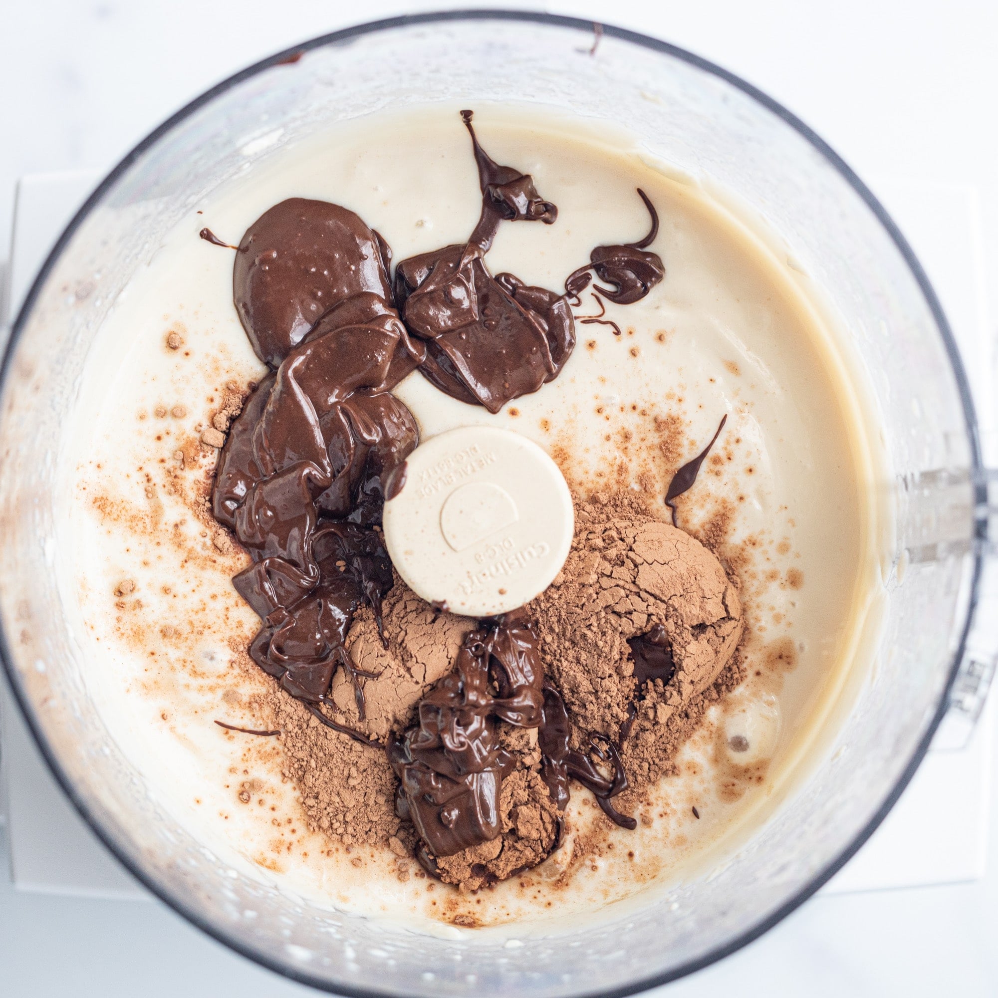 A food processor filled with blended silken tofu, cacao powder, and a drizzle of melted chocolate.