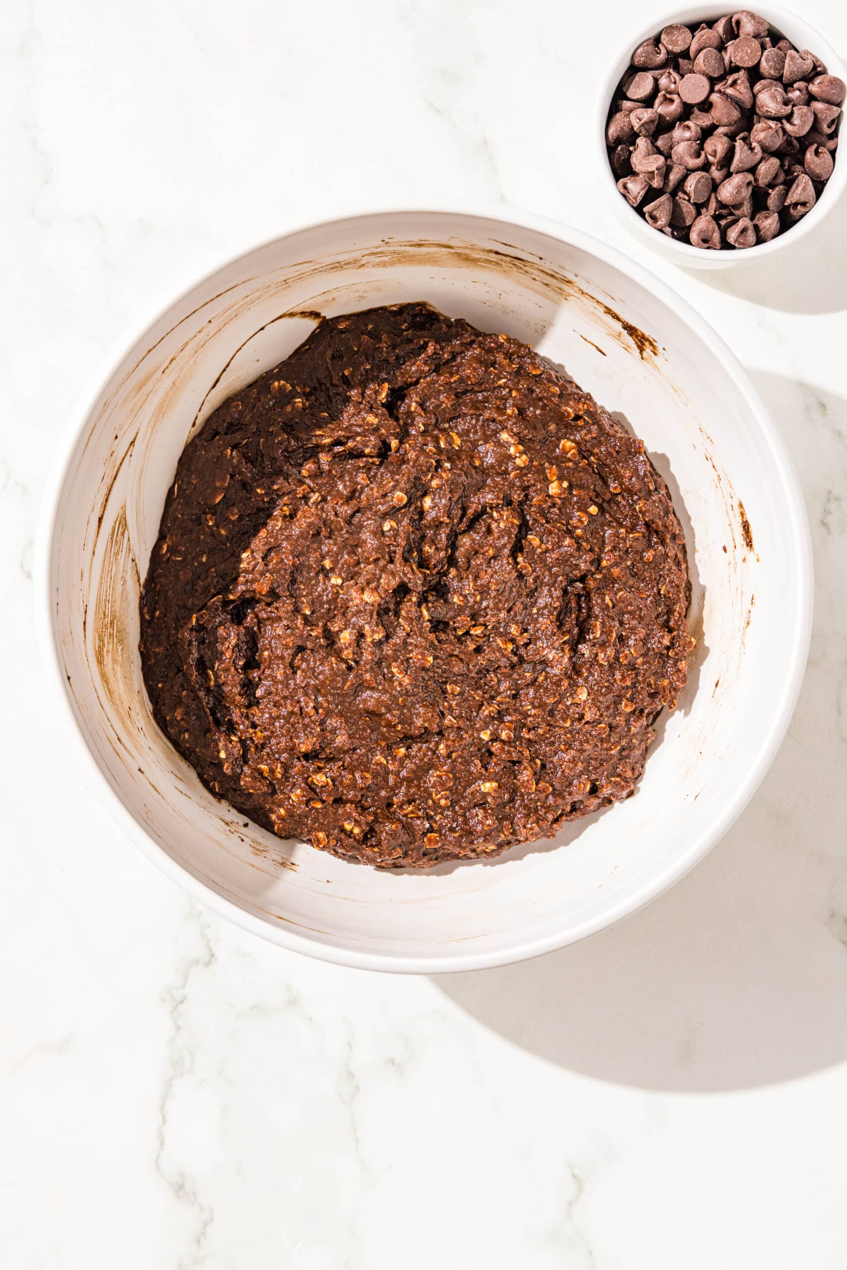A large white bowl of chocolate peanut butter banana muffin batter in it and a smaller white bowl next to it with chocolate chips in it.