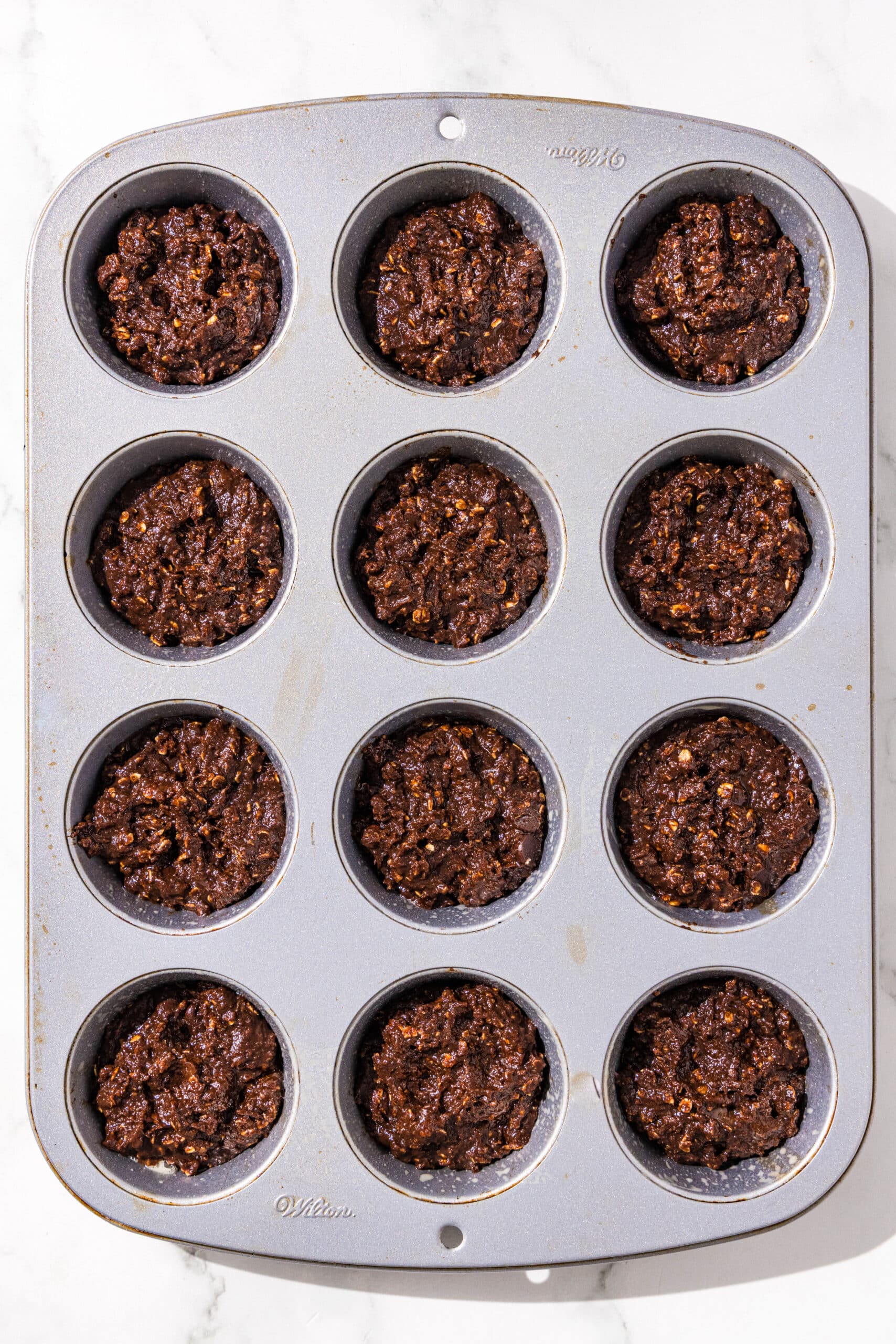 A silver muffin tin filled with 12 chocolate peanut butter banana muffins ready to bake.