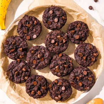 A white plate with a piece of parchment paper on it and 12 chocolate peanut butter banana muffins on it. Chocolate chips, a ripe banana, and a black spoon with peanut butter on it surround the plate.