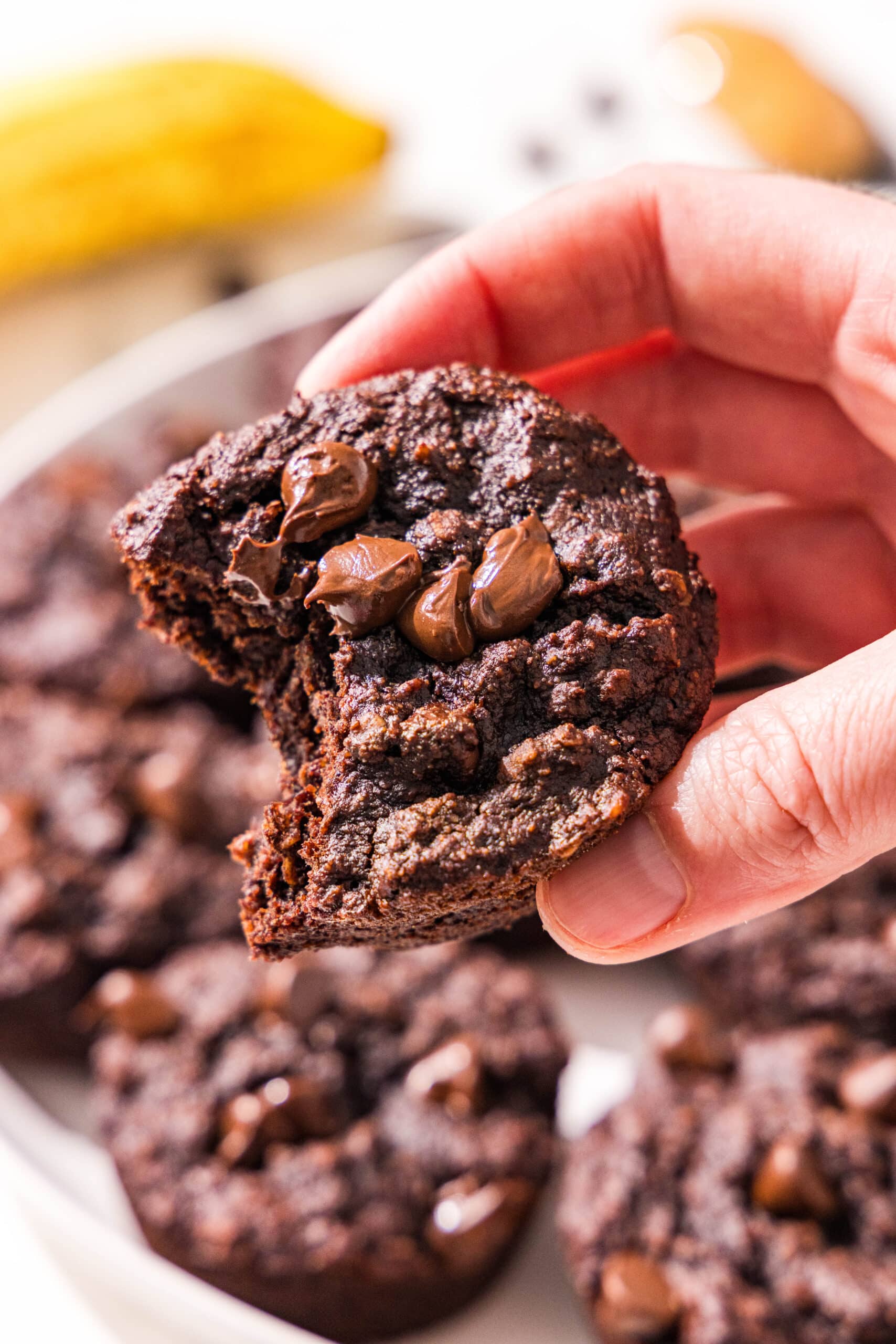 A white hand holding a chocolate peanut butter banana muffin with a bite out of it, over a plate of the muffins. A ripe banana is in the background.