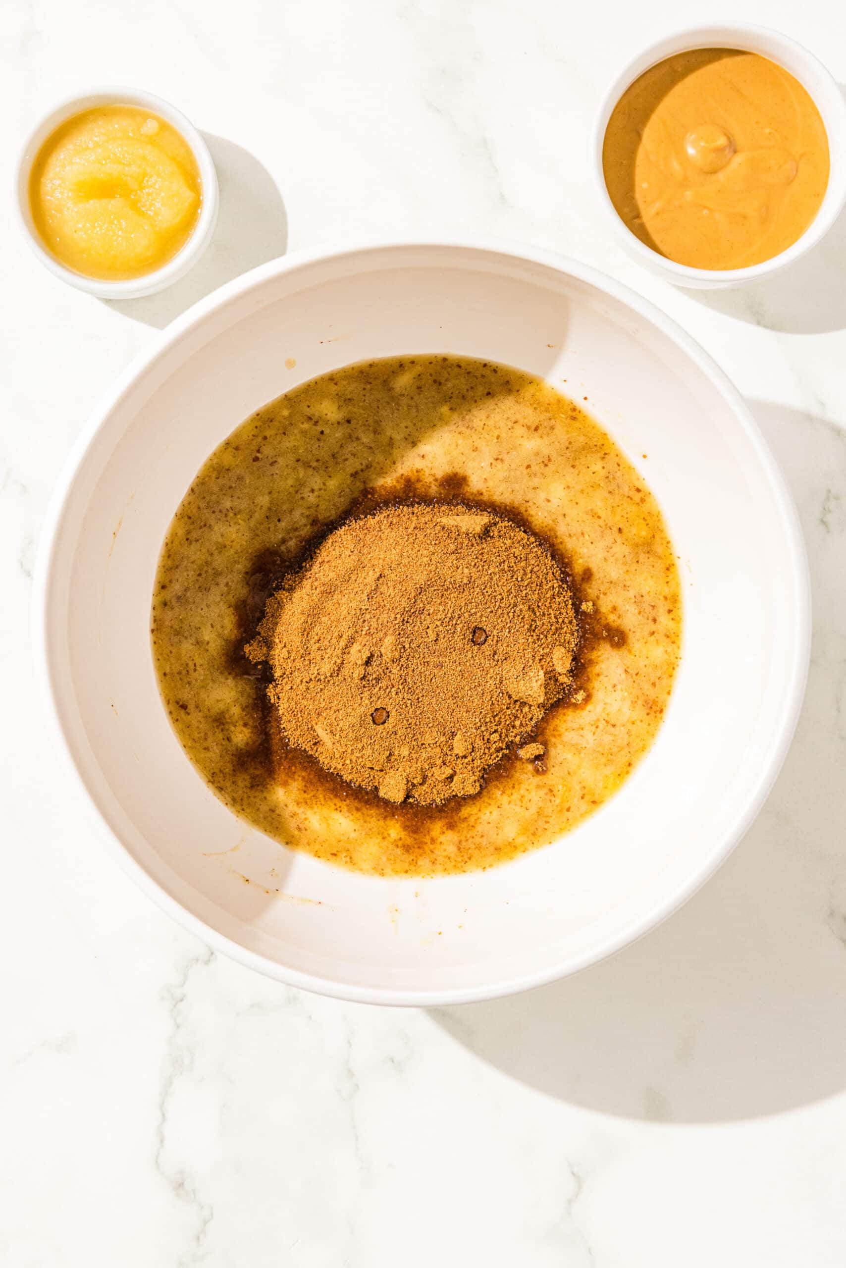 A large white bowl with mashed banana and a flax egg mixed together with a scoop of coconut sugar in the center. Above it is two small white bowls of peanut butter and apple sauce.