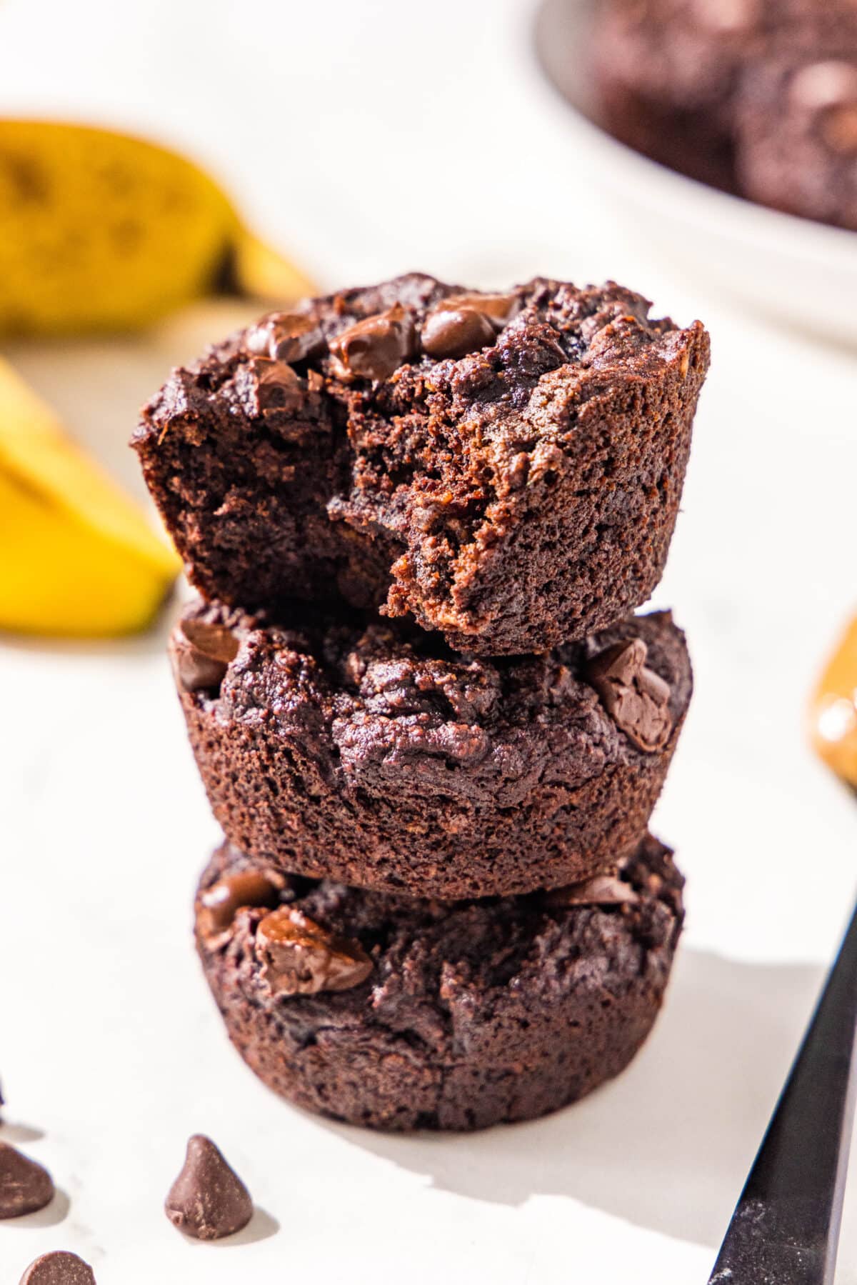Three chocolate peanut butter banana muffins on a white plate with two bananas in the back and a couple chocolate chips to the left.