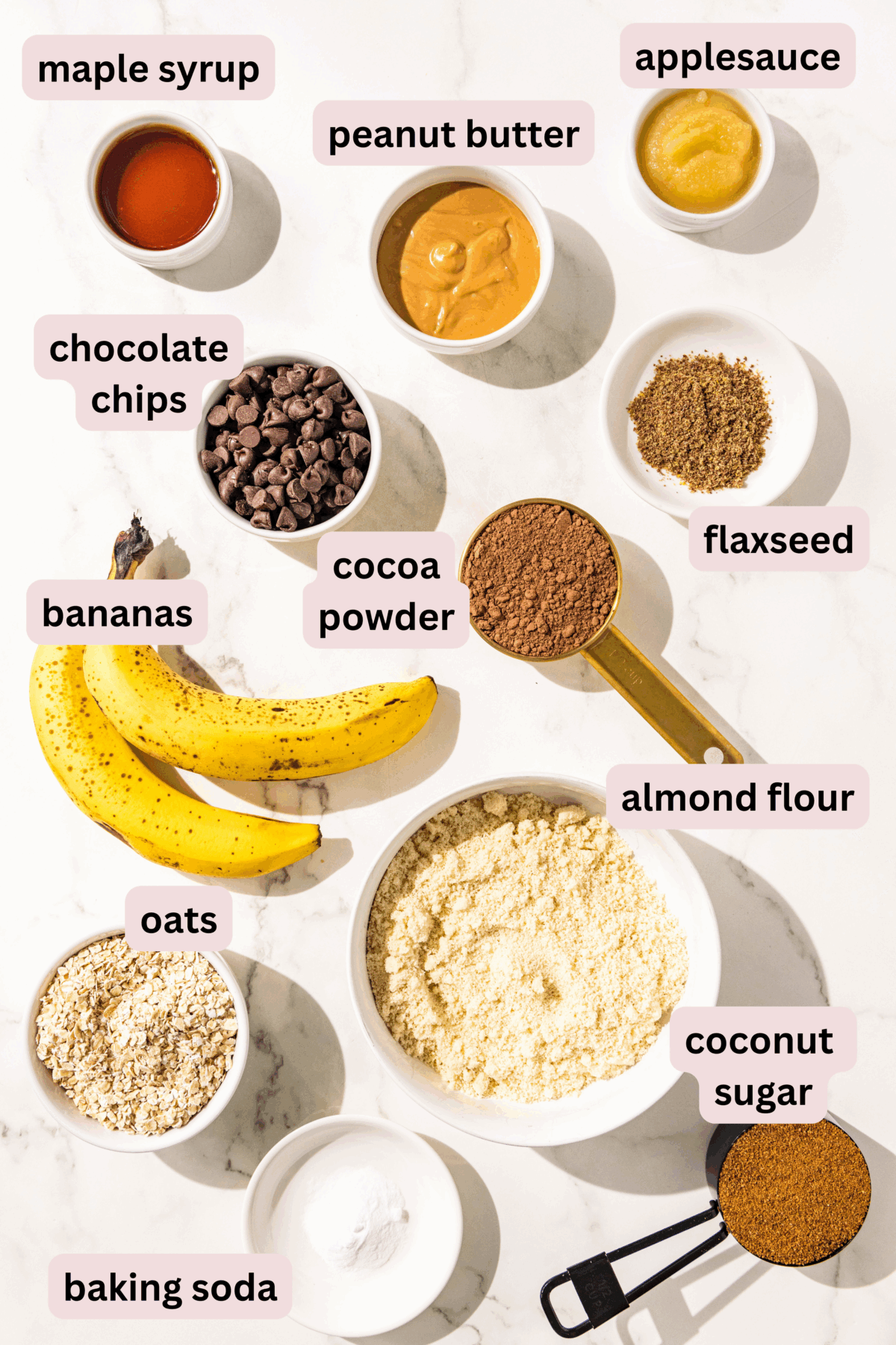 Ingredients needed to make chocolate peanut butter banana muffins laid out on a marble counter including: coconut sugar, baking soda, almond flour, oats, ripe banana, cocoa powder, flaxseed, chocolate chips, peanut butter, apple sauce, and maple syrup. 