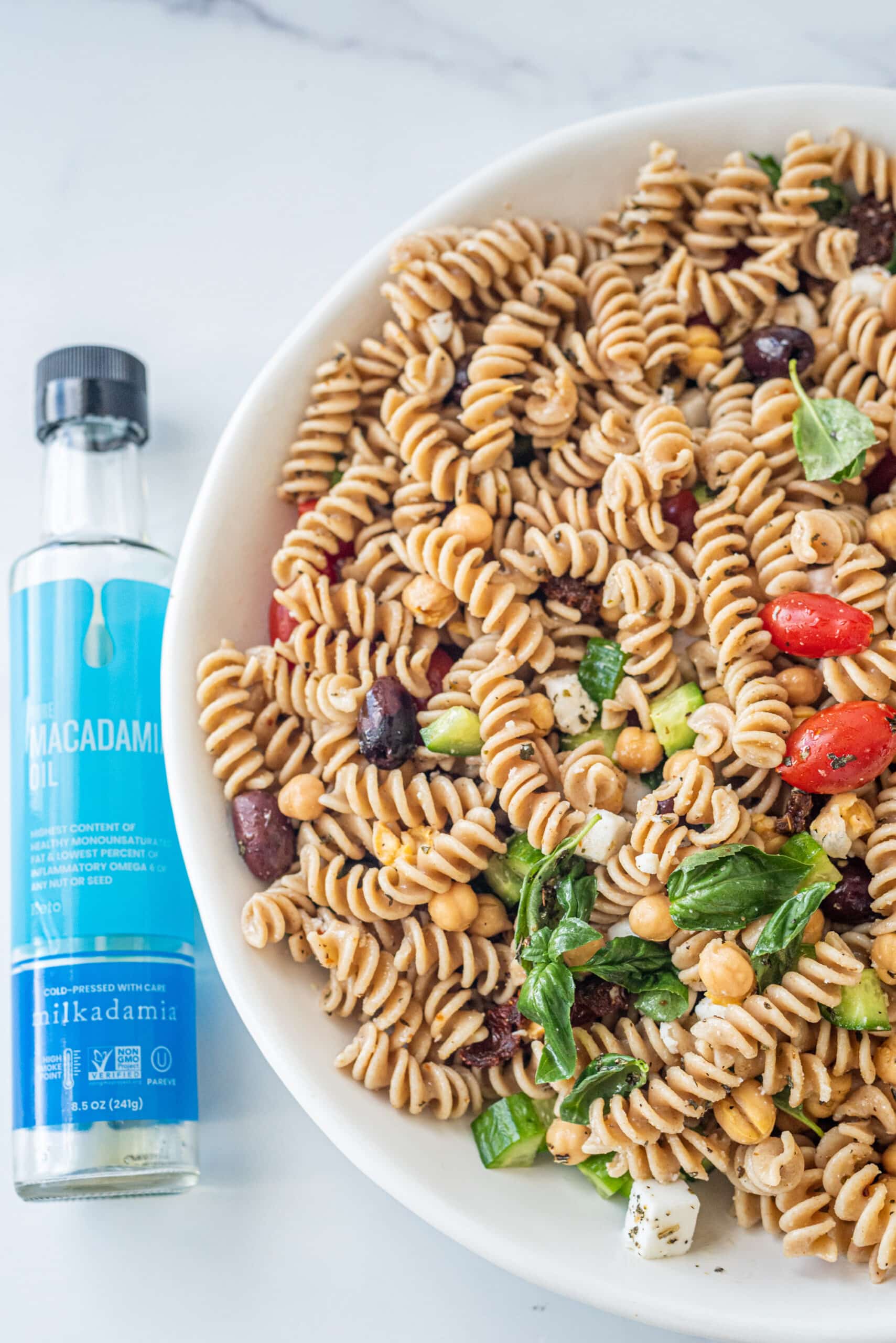 A large white bowl of tossed Vegan Greek Pasta Salad with a clear bottle to the left of macadamia oil.