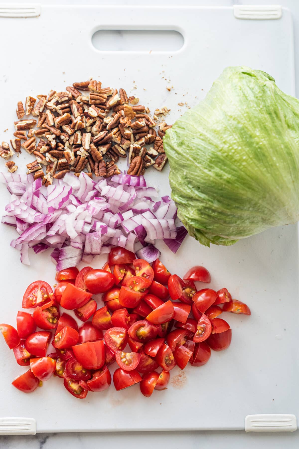 A cutting board with 4 sections of ingredients chopped including pecans, red onion, cherry tomatoes, and iceberg lettuce.