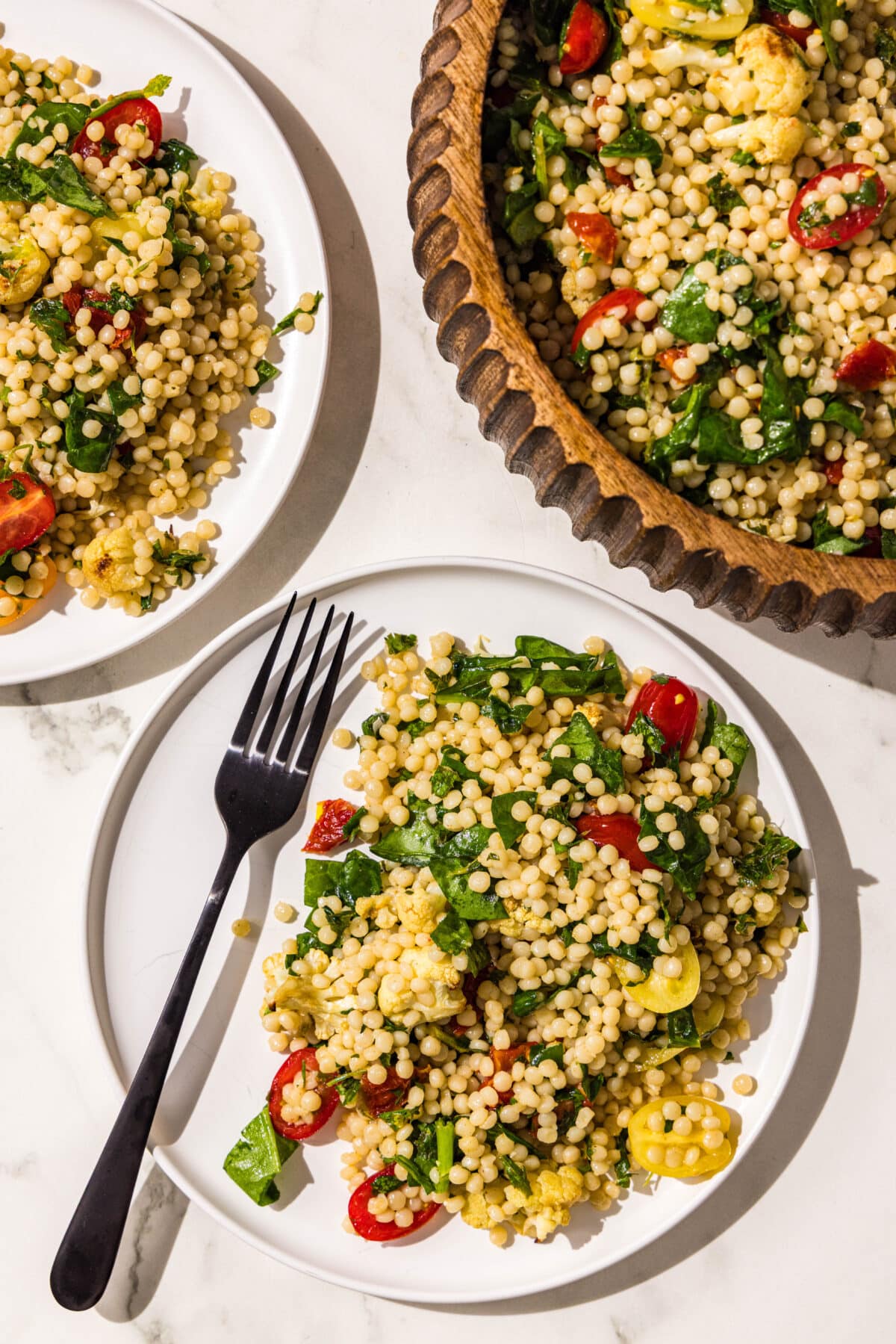Two white plates with a serving of vegan mediterranean couscous and one of them has a black fork on it. To the right of the plates is a large wooden bowl filled with the remaining salad.