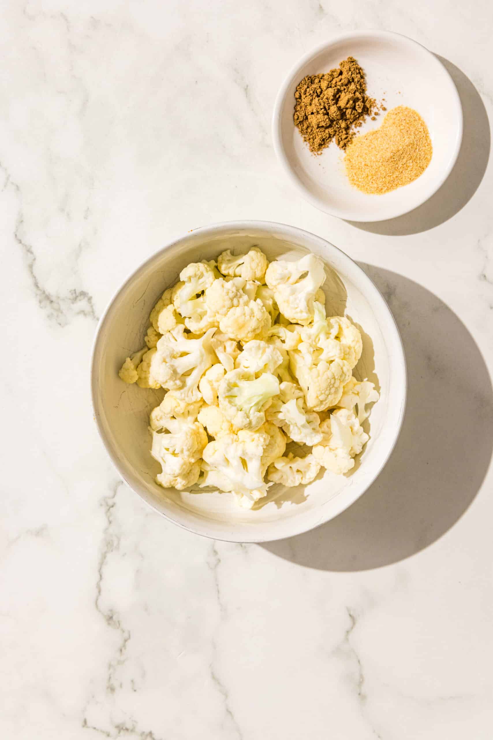A white bowl of raw chopped cauliflower and a smaller white bowl filled with garlic powder and cumin powder.