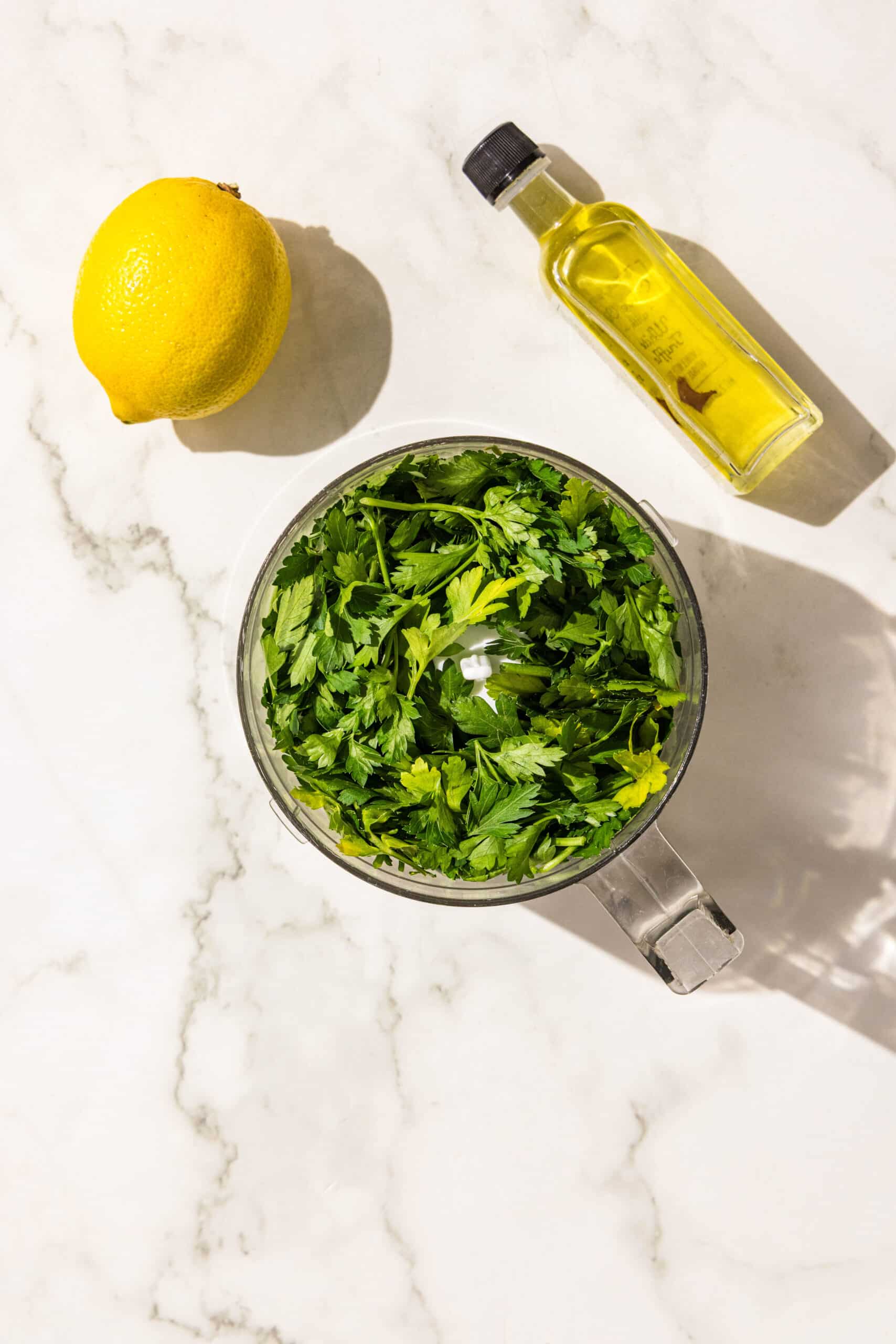 A small food processor with parsley in it. Above it is a lemon and a small bottle of olive oil.