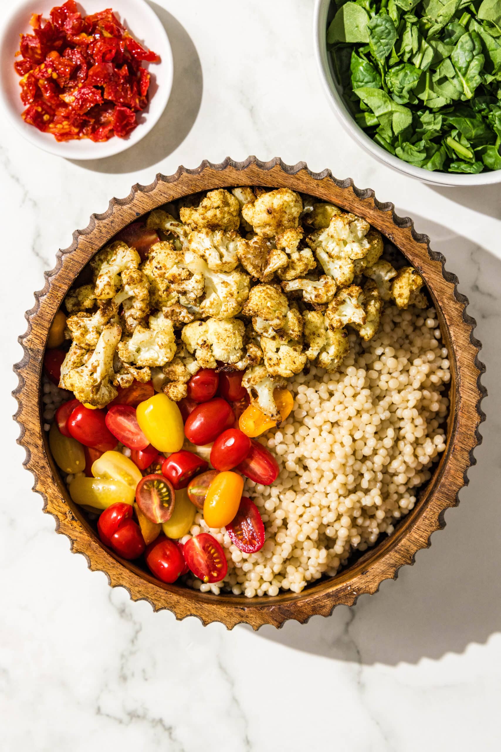 A large wooden bowl of couscous, cherry tomatoes, and roasted cauliflower. Above it is two smaller white glass bowls of sundried tomatoes and chopped spinach.