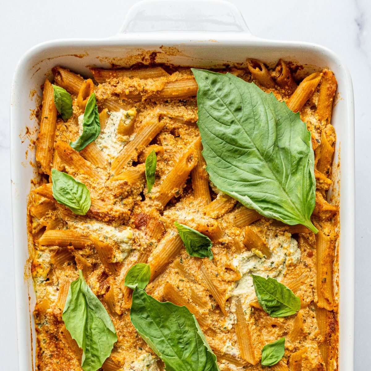 A white casserole dish of dairy-free baked ricotta pasta topped with fresh basil.