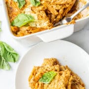 A casserole dish filled with baked pasta topped with fresh basil, a fork in the dish, and a small white plate with a scoop of pasta.