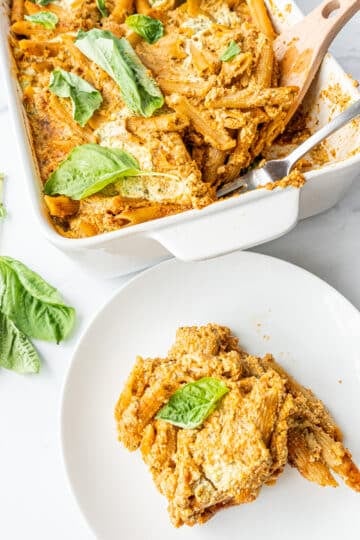 A casserole dish filled with baked pasta topped with fresh basil, a fork in the dish, and a small white plate with a scoop of pasta.