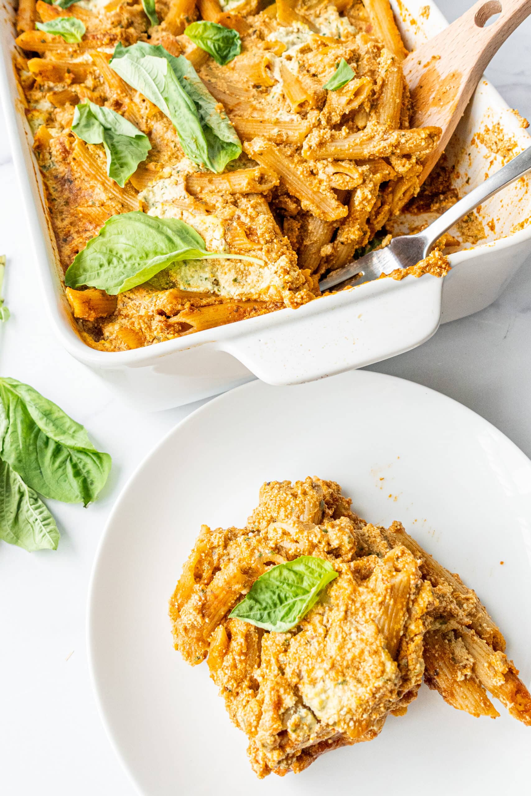 A casserole dish filled with baked pasta topped with fresh basil, a fork in the dish, and a small white plate with a scoop of pasta.