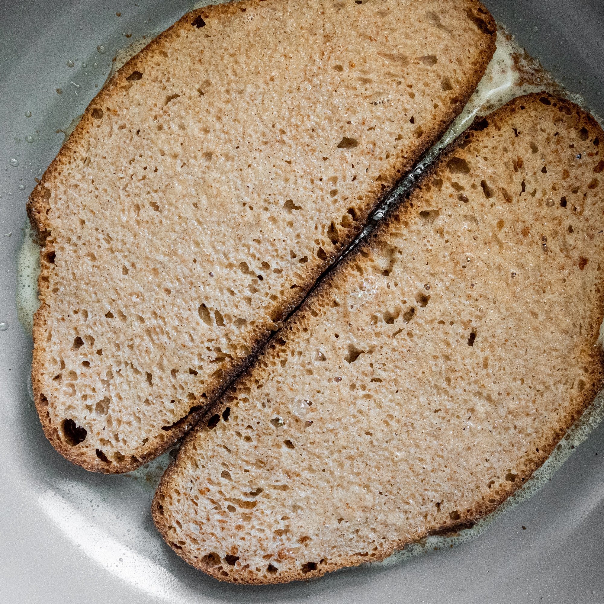 Two pieces of sourdough bread that have been dipped in french toast batter laid side by side in a grey frying pan.